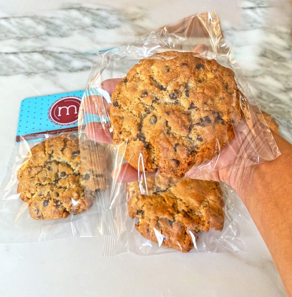 Una mano sostiene una galleta con chispas de chocolate que está guardada en una bolsa transparente. En el fondo de la imagen se observan dos galletas guardadas en bolsas transparente y sobre ellas una tarjeta con el logo de la repostería "m de motivos".