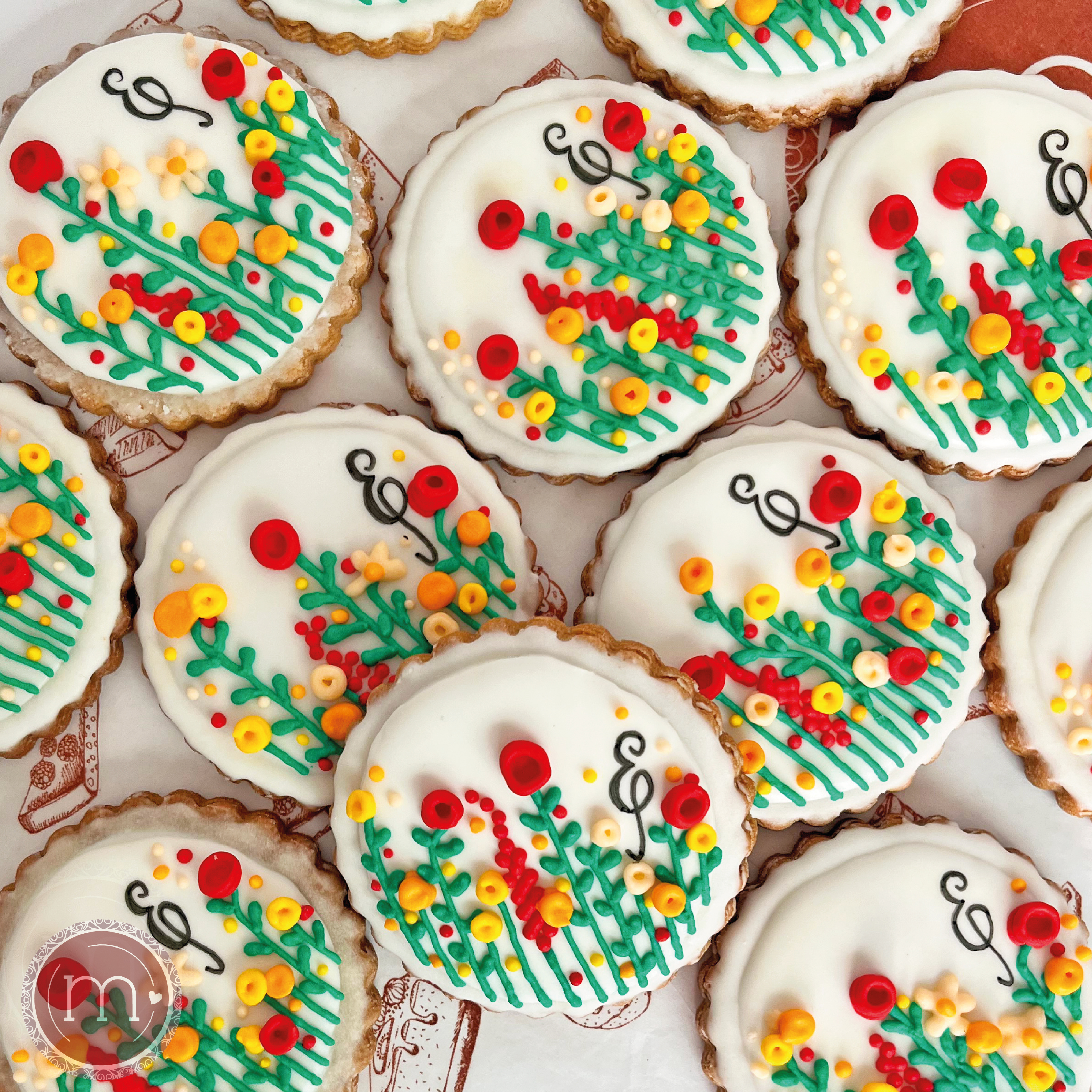 Un grupo de galetas personalizadas con una decoración de flores rojas y amarillas. Todas tienes escrita la letra "E". Las galletas se colocan sobre un papel decorado.