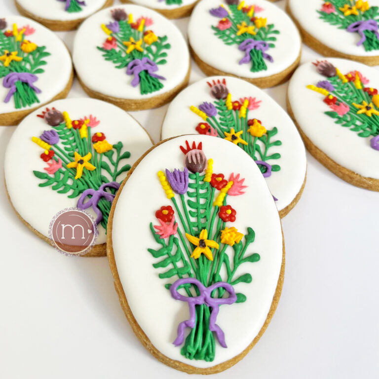 Un grupo de galletas con forma ovalada, decoradas con glaseado blanco y ramos de flores igual hechos con royal icing. Algunas galletas están acostadas y en primer plano se coloca una paada recargada sobre las que están acostadas. Estas galletas personalizadas fueron hechas para una boda..