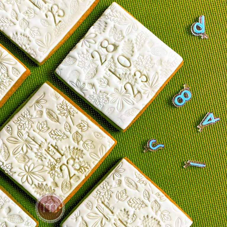 Un grupo de galletas cuadradas decoradas con un glaseado de color blanco y un grabado de flores y números que indican una fecha. Estas galletas personalizadas para boda Tienen grabdas la fecha del evento y nombres. Las galletas se colocan sobre una base de color verde.