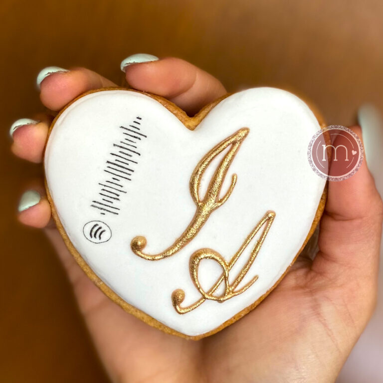 Una galleta en forma de corazón es sostenida por una mano. La galleta tiene una decoración de glaseado blanco y las iniciales "J y A" con letras doradas. Las galletas personalizadas como esta, que tiene dibujado un código escaneable son populares para bodas.
