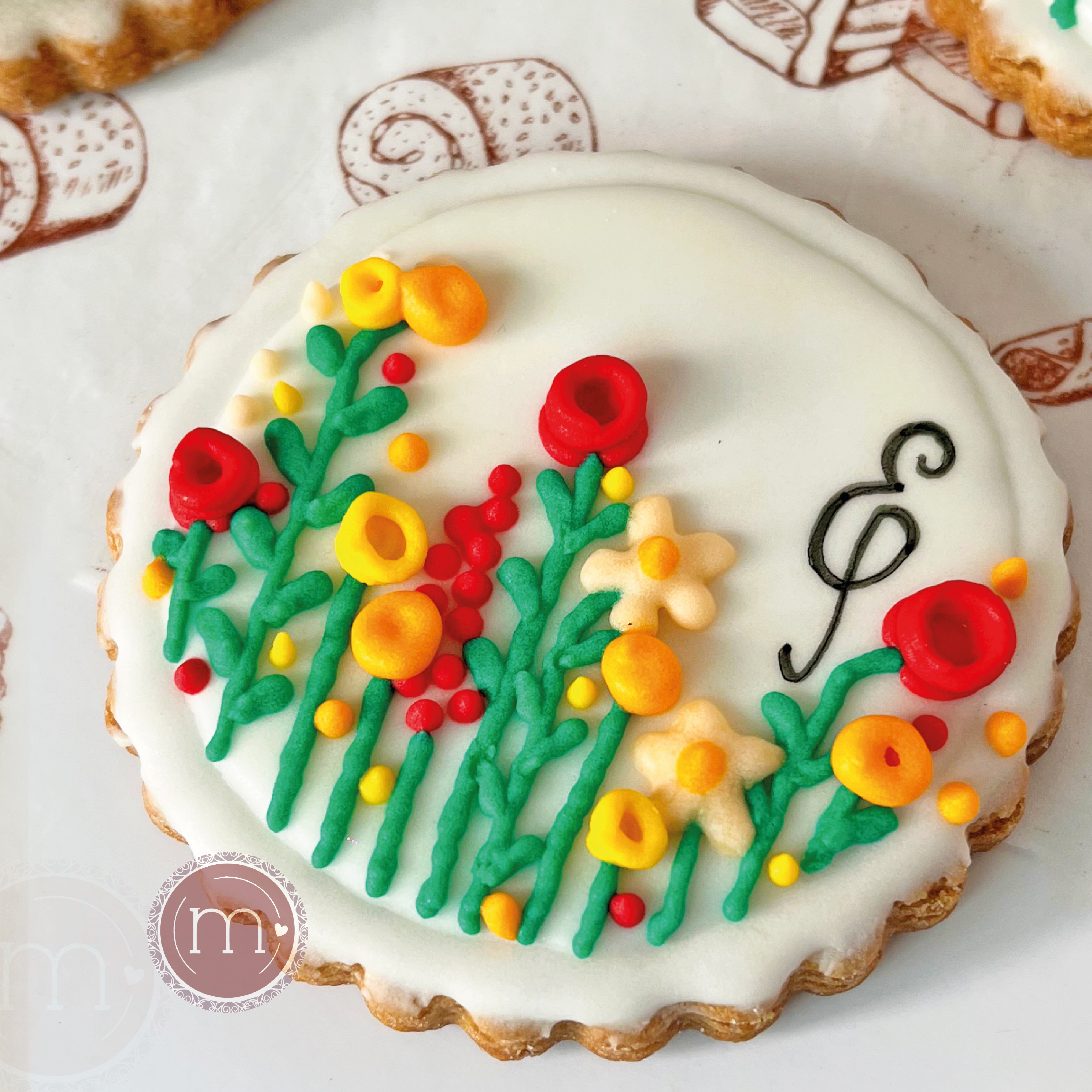 Una galleta decorada con glaseado blanco y un ramo de flores de royal icing. Sobre el glaseado blanco se escribe la letra "E". Estas galletas personalizadas fueron hechas para un baby shower. La galleta se coloca sobre un papel decorado.