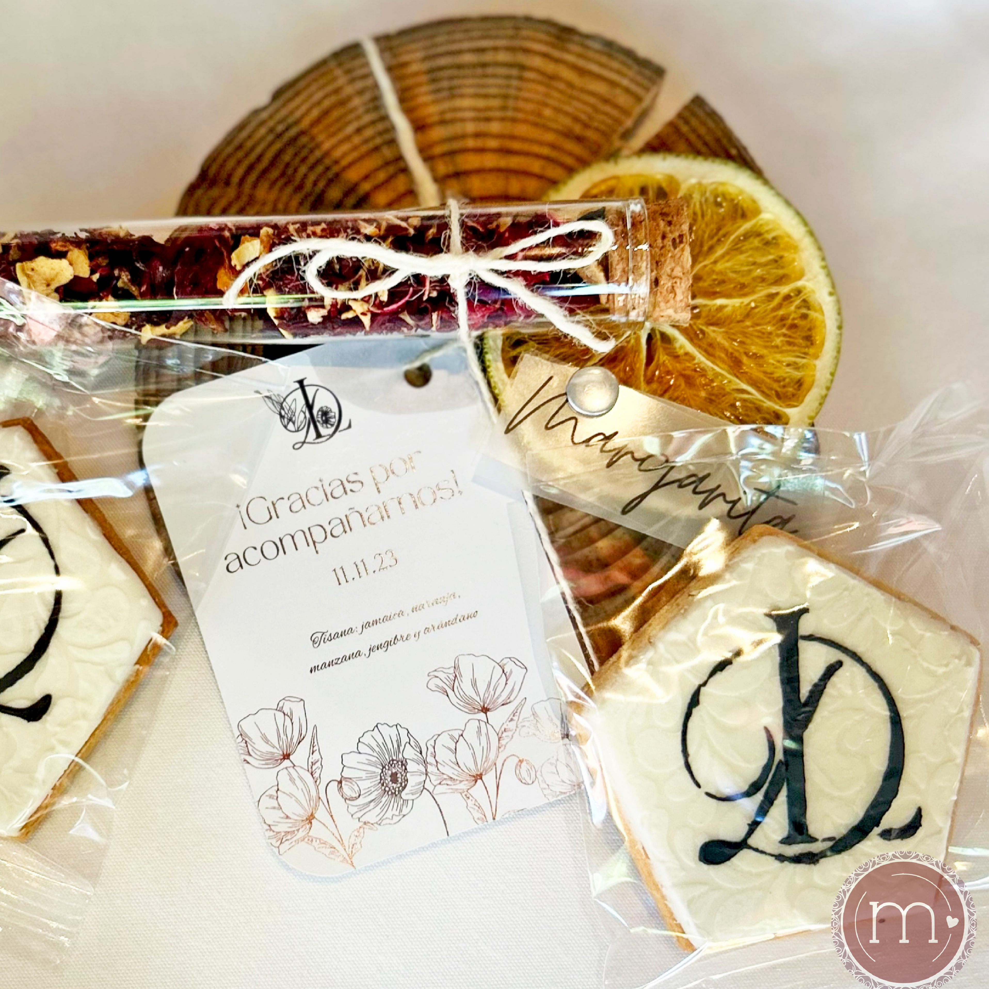 Una galleta decorada con glaseado blanco y con la letra "D" en color negro, guardada dentro de una bolsa transparente. Junto a la galleta se coloca una invitación de boda y un envase con frutos secos. Estas galletas personalizadas son hechas para una boda. La galleta se coloca sobre una base de madera.