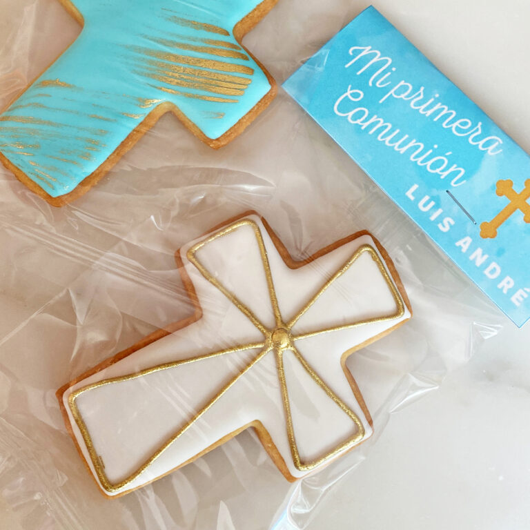 Una galleta en forma de cruz, decorada con un glaseado blanco y líneas doradas. A un lado se coloca una galleta en forma de cruz de color azul. Estas galletas personalizadas para primera comunión se colocan dentro de una bolsa transparente con una etiqueta que dice "primera comunión.
