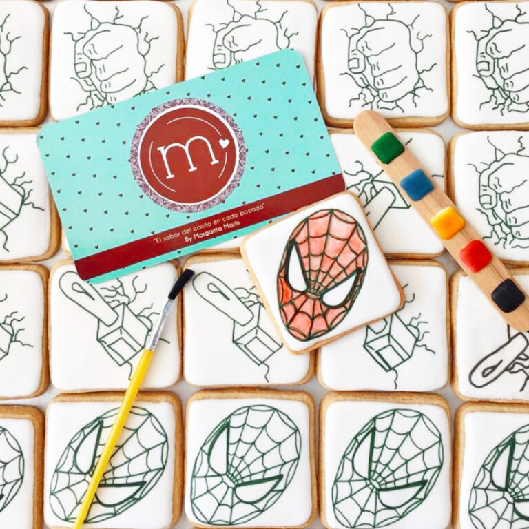 Una galleta decorada con un glaseado blanco y un dibujo del personaje "spiderman", junto a ella se coloca un pincel, una tira de colores y una tarjeta azul con el logo de la repostería "m de motivos". Debajo se coloca un grupo de galletas con un trazo de la cara de "spiderman". Estas galletas personalizadas fueron hechas para una fiesta infantil.