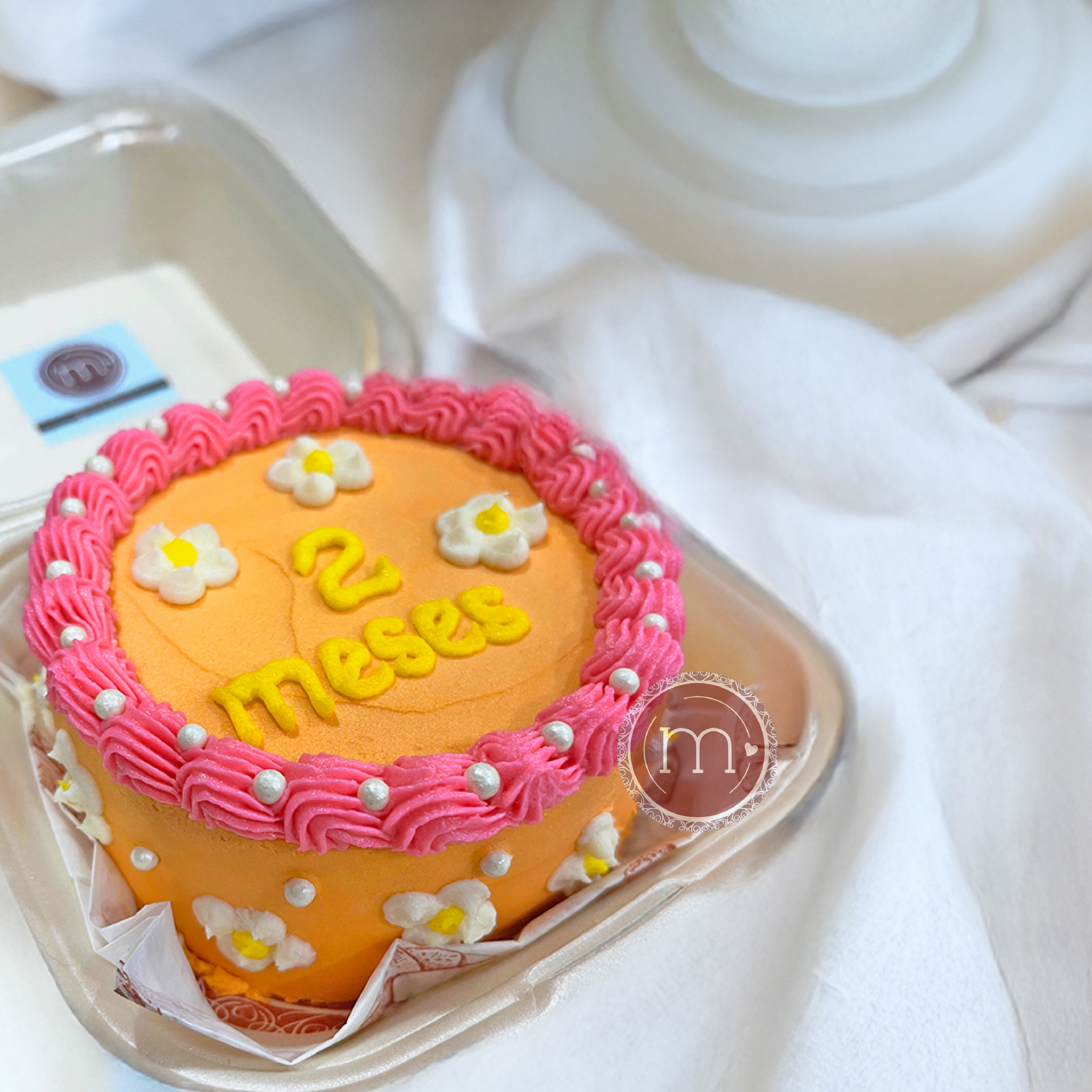 Un pastel decorado con betún naranja y rosa. Este mini pastel personalizado tiene la frase "2 months" escrita con letras de color amarillo y perlas. El pastel se coloca dentro de un recipiente blanco con papel y a su vez sobre un mantel de tela color blanco.