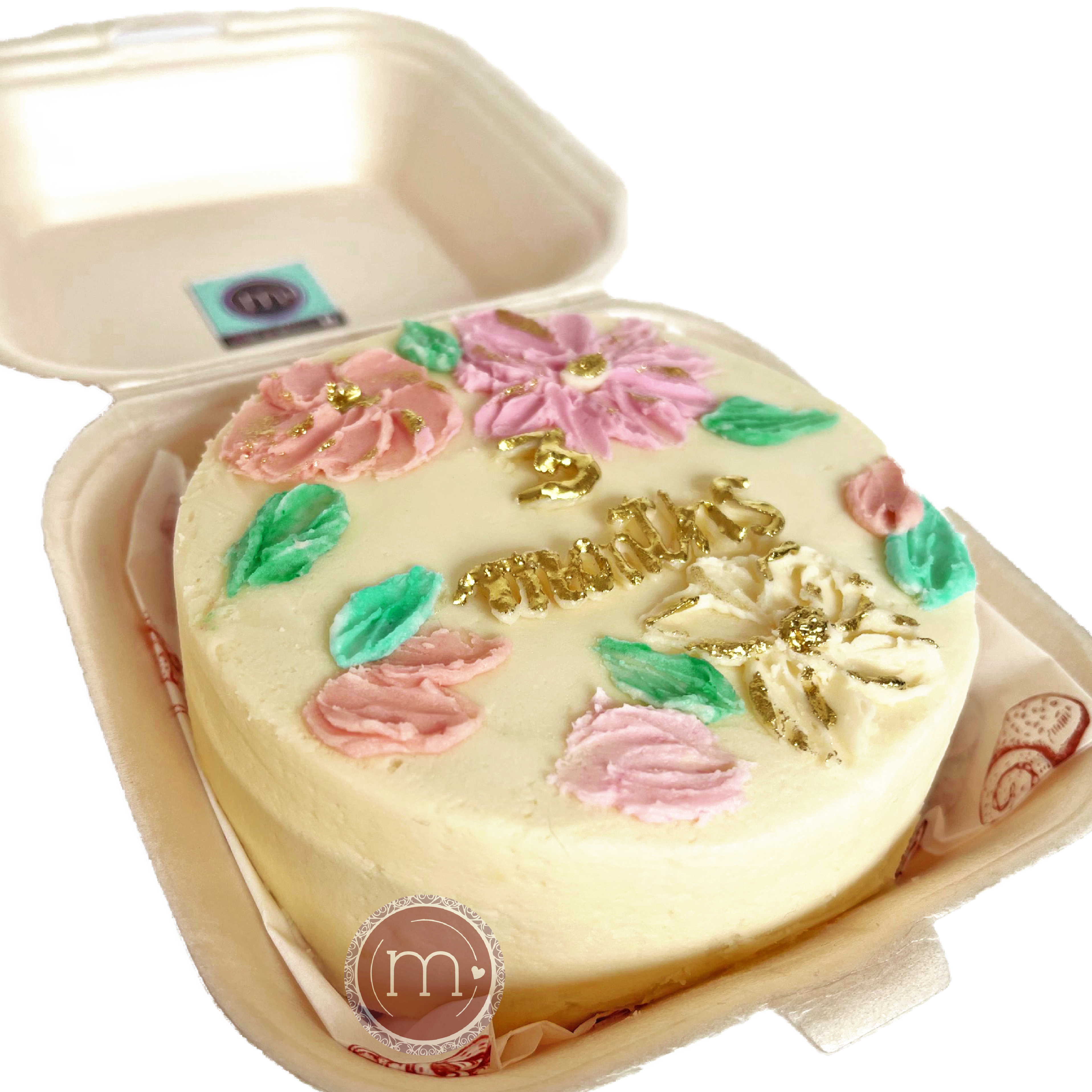 Un mini pastel personalizado con la palabra "3 months" de letras doradas. El pastel está decorado con betún de color blanco y petalos de betún de color verde y lila. El pastel se coloca dentreo de un recipiente blanc .
