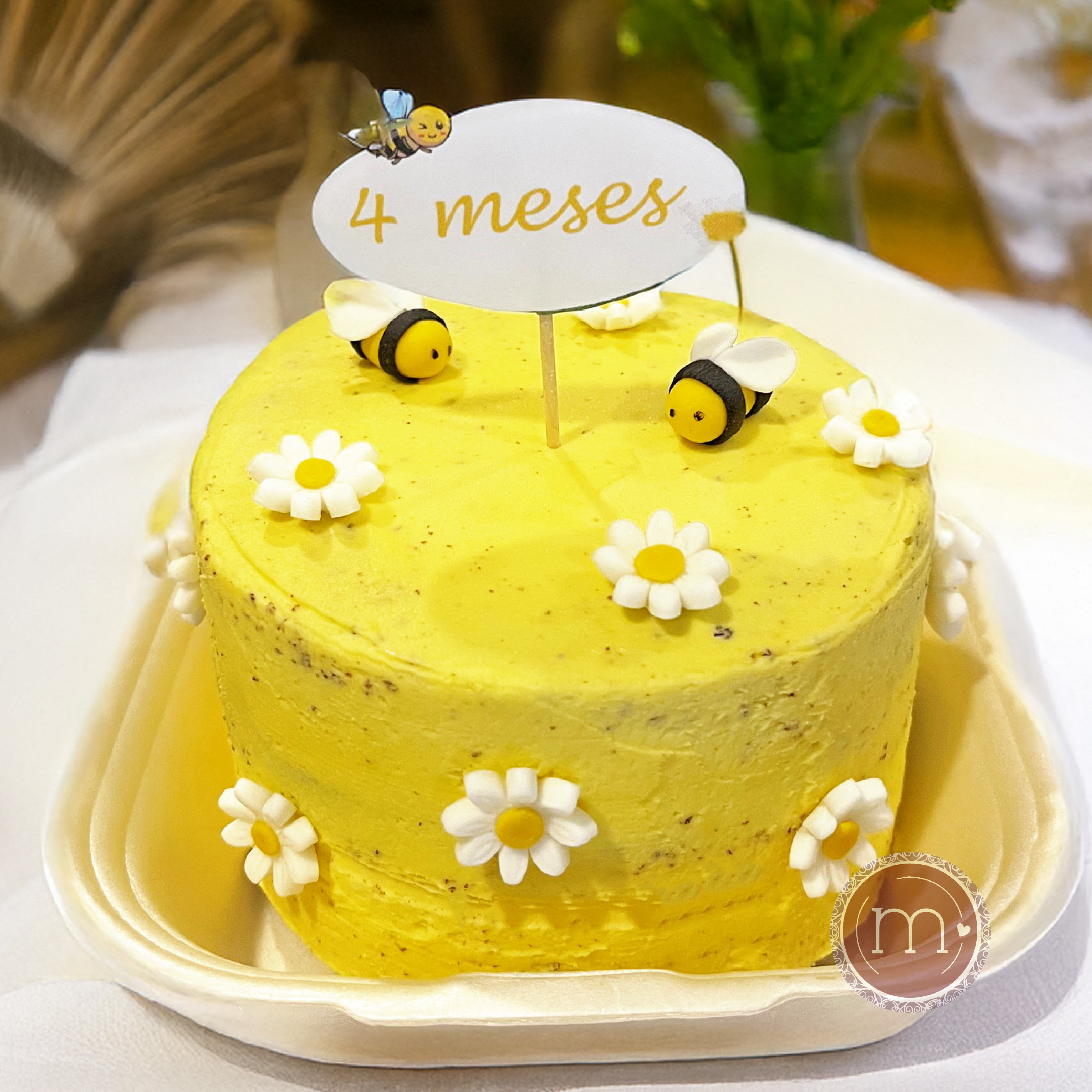 Un pastel decorado con betún de color amarillo y con pequeñas flores blancas de fondan y abejas hechas de fondant. Este mini pastel personalizado para una sesión de fotos smash cake tiene un letrero que dice "4 meses". El pastel se coloca dentro de un recipiente blanco y sobre un mantel de tela color blanco.