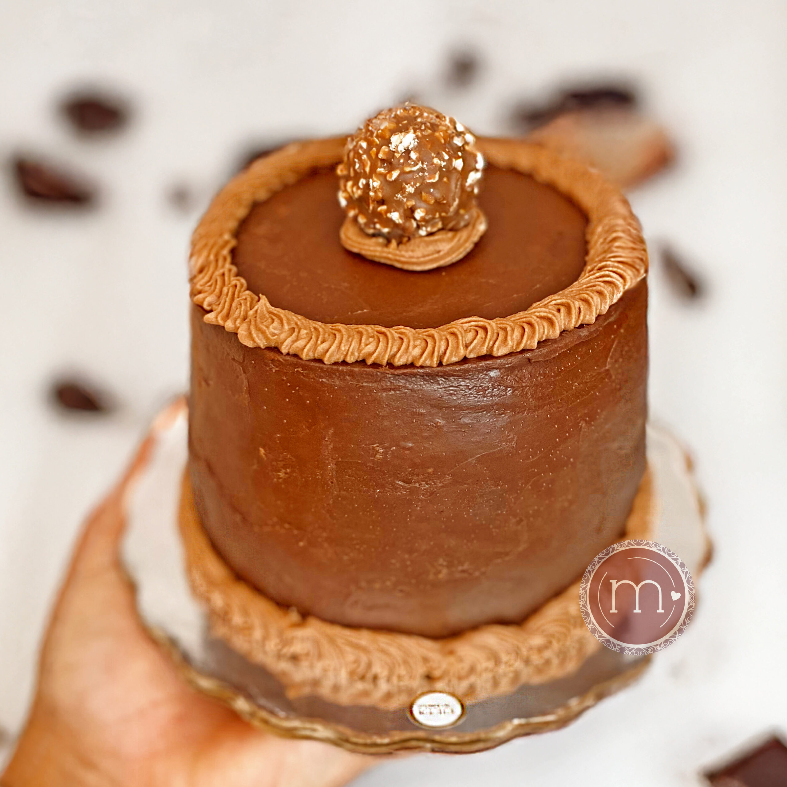 Un mini pastel personalizado de sabor chocolate ferrero. En la parte superior se coloca un chocolate "Ferrero". El pastel es sostenido por una mano.