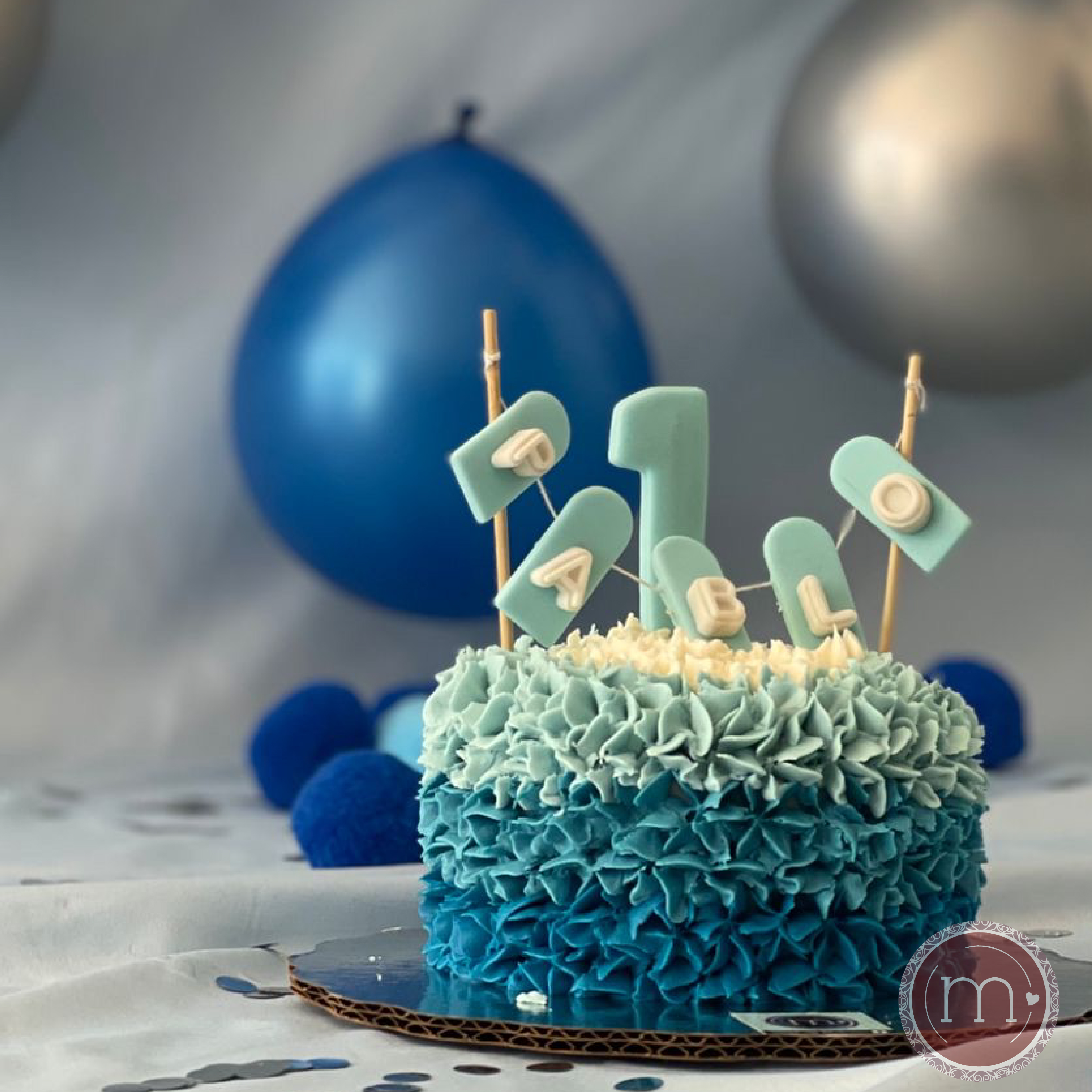 Un mini pastel personalizado con betún de color azul en degradado. En la parte superior se coloca un letrero de fondant que dice "Pablo" y el número "1". Este mini pastel es para una sesión de fotos de un bebé que cumple 1 año. El pastel se coloca sobre una base reflectante y de fondo se observan globos azules y grises.