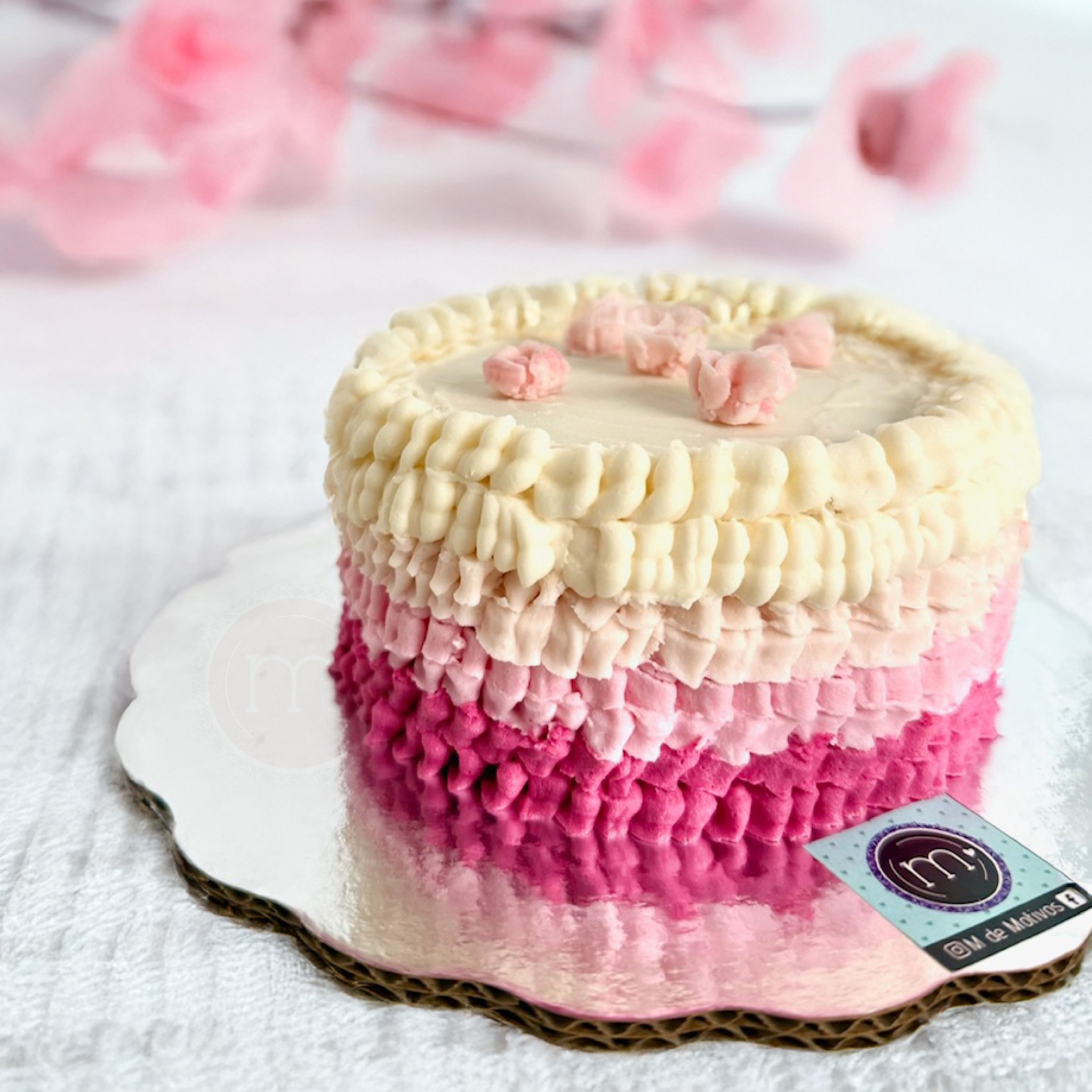 Un mini pastel decorado con betún en dredado del color rrosa. El pastel se colca sobre una base plateada reflectante con una etqieuta que tiene el logo de postres personalizados m de motivos.