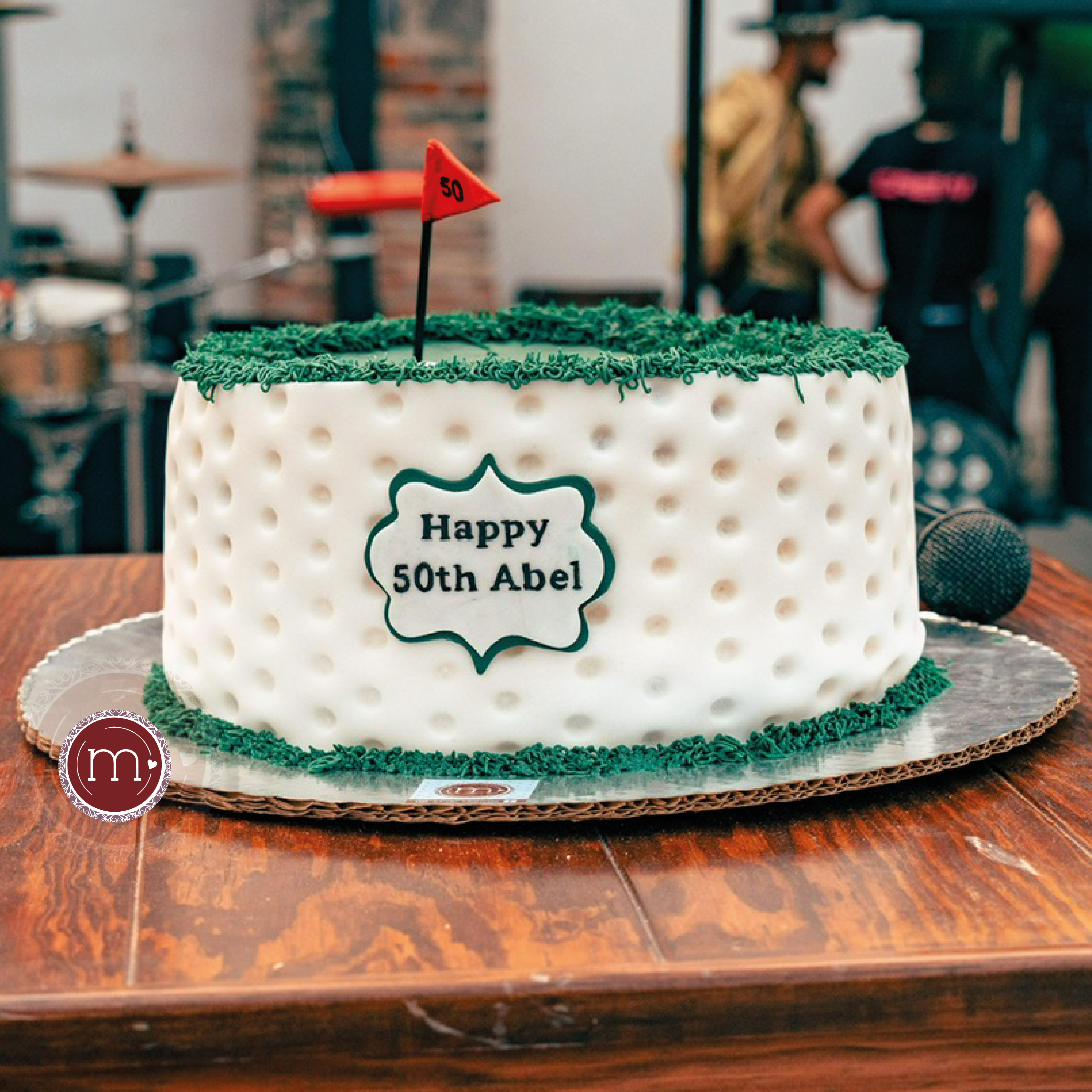 Un pastel redondo decorado con fondant con temática de golf. En la parte frontal tiene una placa que dice "Happy Birthday". Los pasteles fondant son ideales para los eventos temáticos pues son más fáciles de personalizar. El pastel se coloca sobre una base de madera.