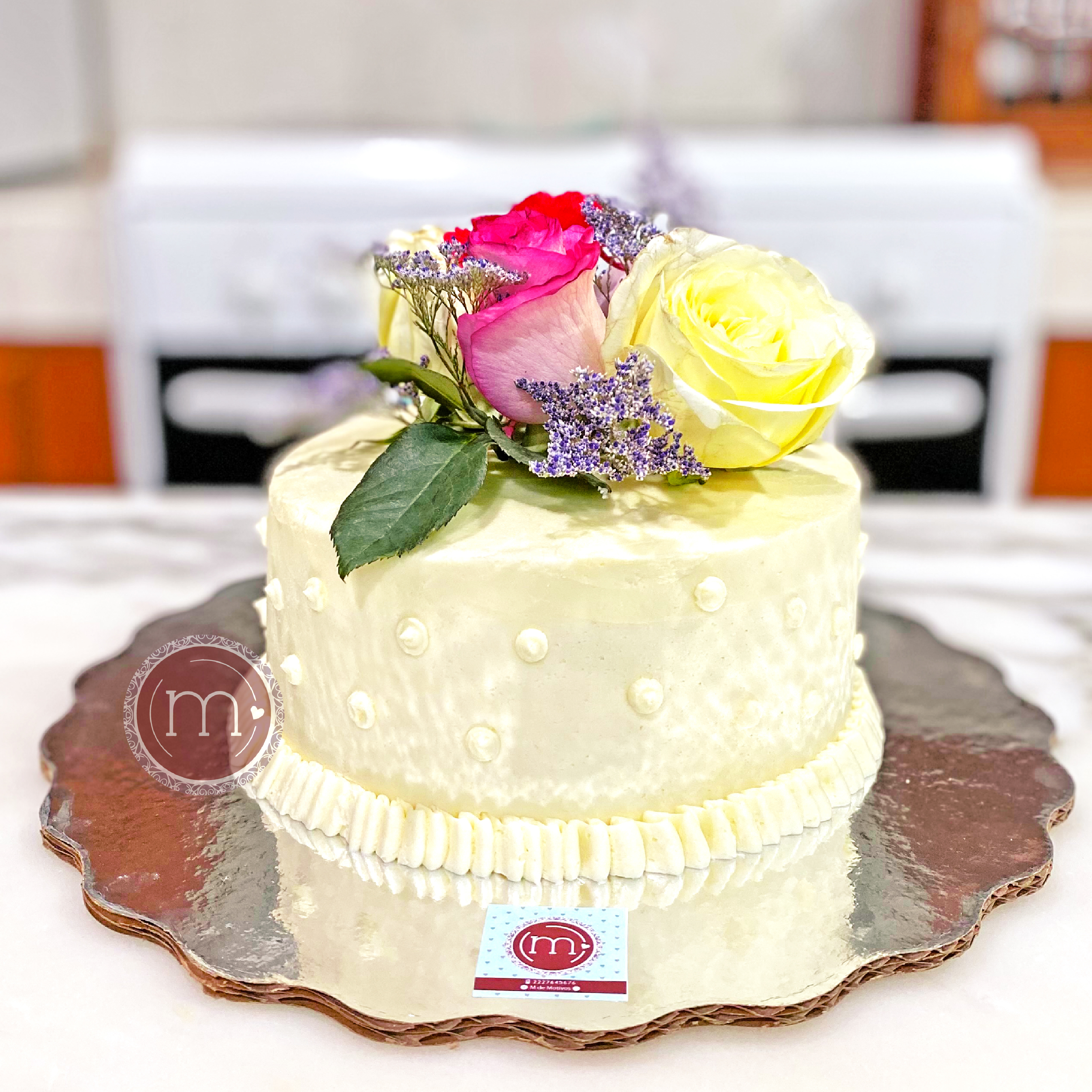 Un pastel pequeño sobre una base de cartón plateado. El pastel está decorado con piedras comestibles blancas y con tres rosas en la parte superior. El pastel se coloca sobre una mesa blanca. Para las personas que buscan un pastel de bodas pequeño, m de motivos tiene este bello diseño.