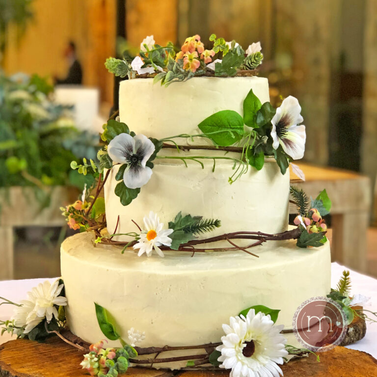 Pastel glaseado blanco de tres niveles adornado con margaritas blancas, colocado sobre una base de troncos de madera. El pastel de bodas personalizado con flores tiene como fondo un exuberante follaje verde y un muro de piedra.