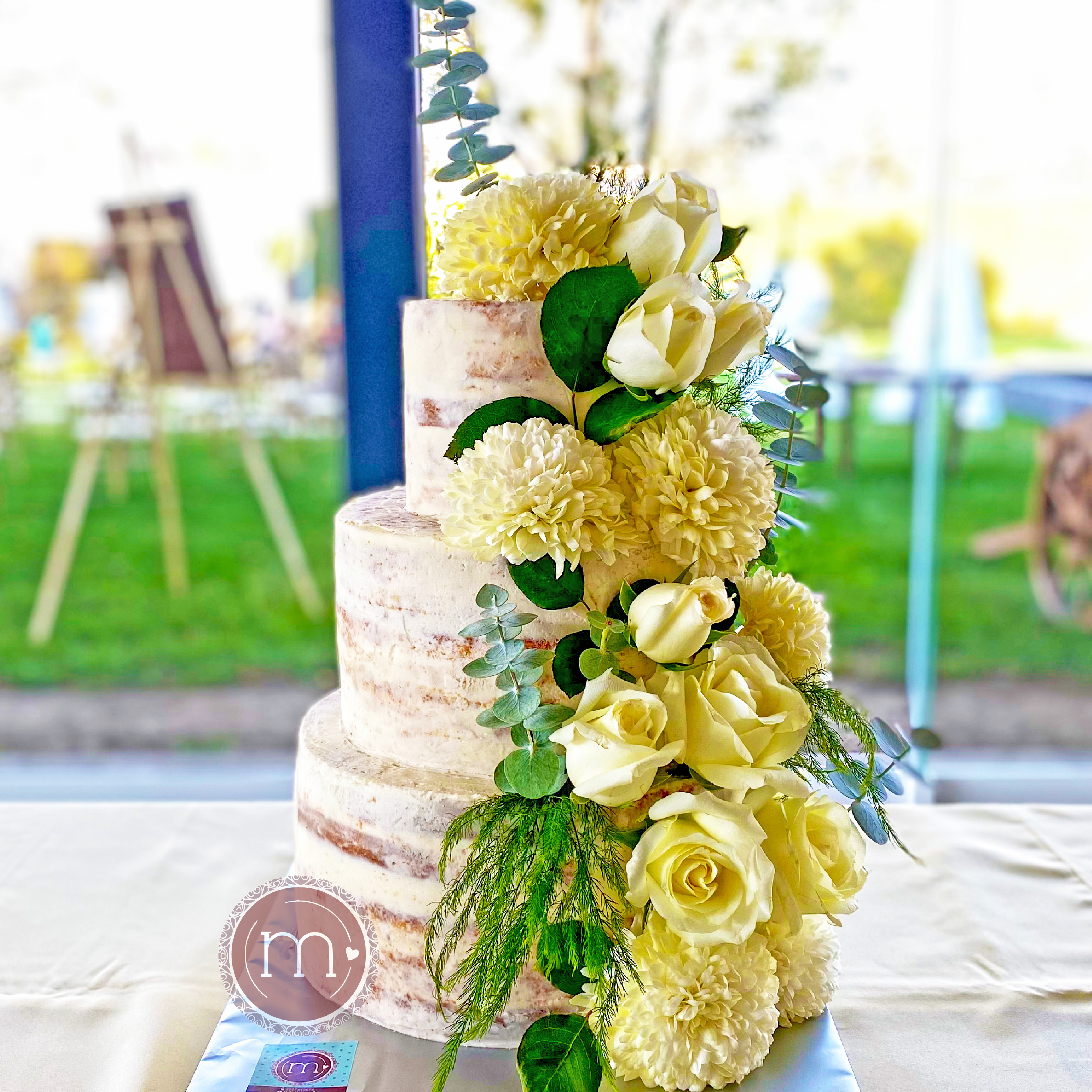 Un pastel de tres pisos con cubierta blanca, está decorado con una tira de flores, rosas blancas y hojas verdes. El pastel está sobre una base de color platado y a su vez sobre una tela blanca. Este estilo de pastel "naked cake" es un diseño ideal de pastel de bodas.
