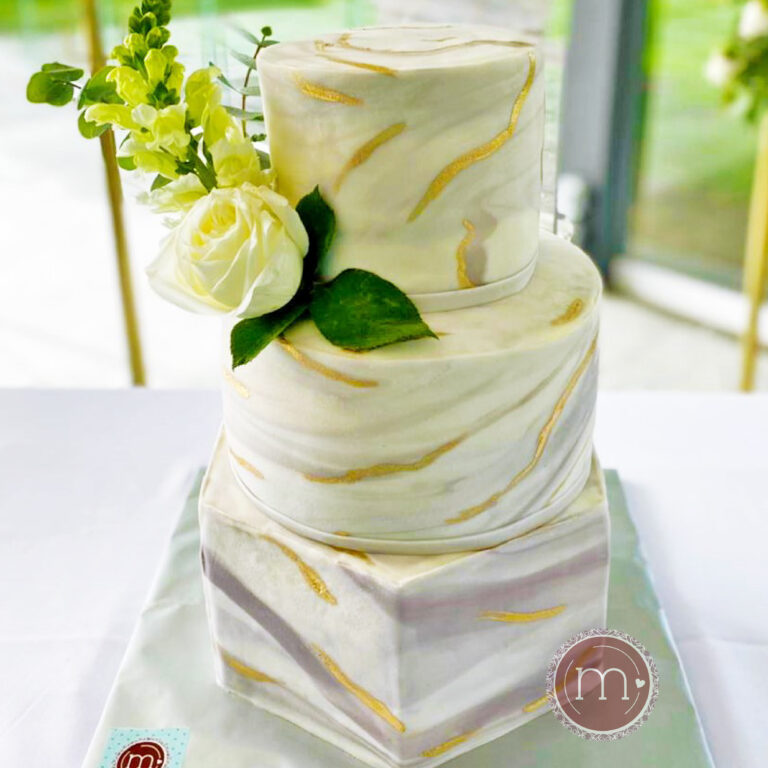 Un pastel de bodas de tres piso decorado con fondant con un diseño tipo mármol. El primer piso es en forma de hexagono, el segundo en forma de circulo y el tercer piso también forma de circulo con un diametro menor al segundo piso y con mayor altura. Entre el segundo y tercer piso se coloca una rosa blanca otras pequeñas flores amarillas. El pastel se coloca sobre una base de color plateado. Al fondo de la imagen se observa un jardín..