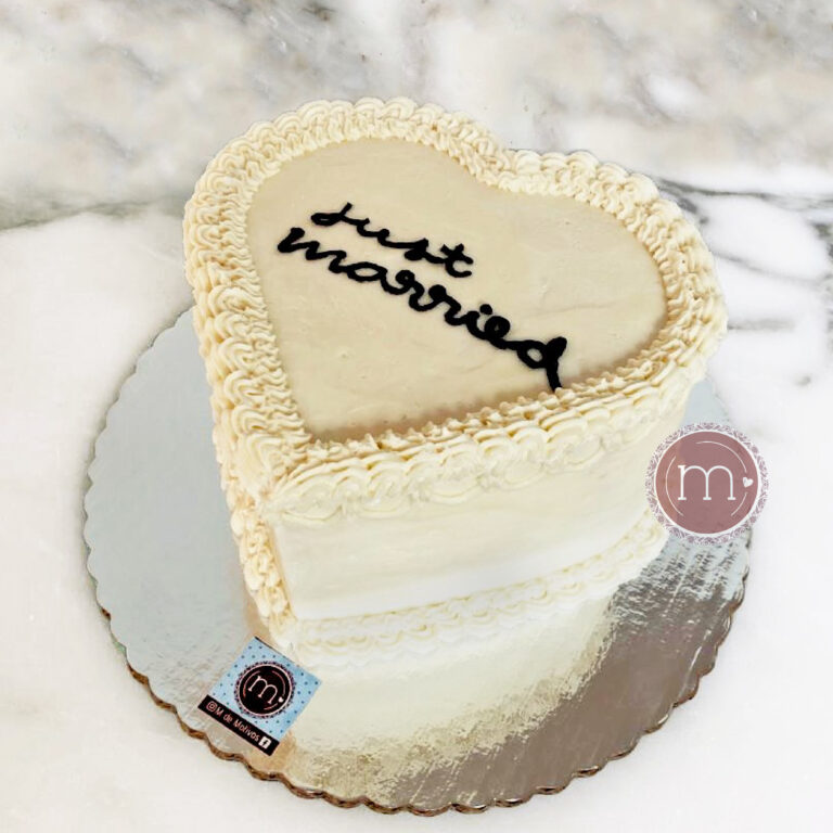 Un pastel de bodas en forma de corazón decorado con betún blanco. En la parte superior del pastel está escrito con letra cursiva en color negro "Just Married". El pastel se coloca base de cartón plateado reflejante.