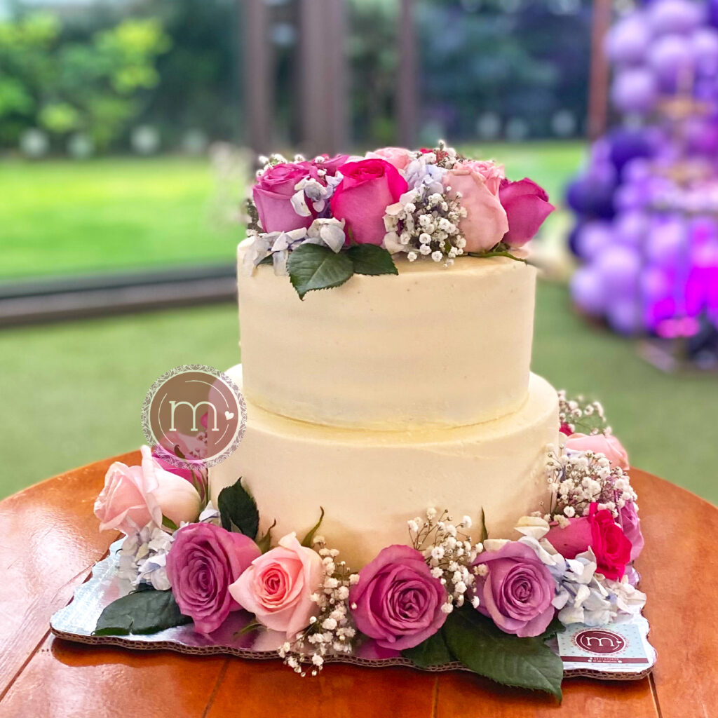 Un pastel de dos pisos decorado con betún blanco y flores alrededor del primer piso y un pequeño ramo sobre el del segundo piso. Este pastel de bodas se coloca sobre una mesa de madera. Detrás del pastel se observa un jardín con decoración morado.