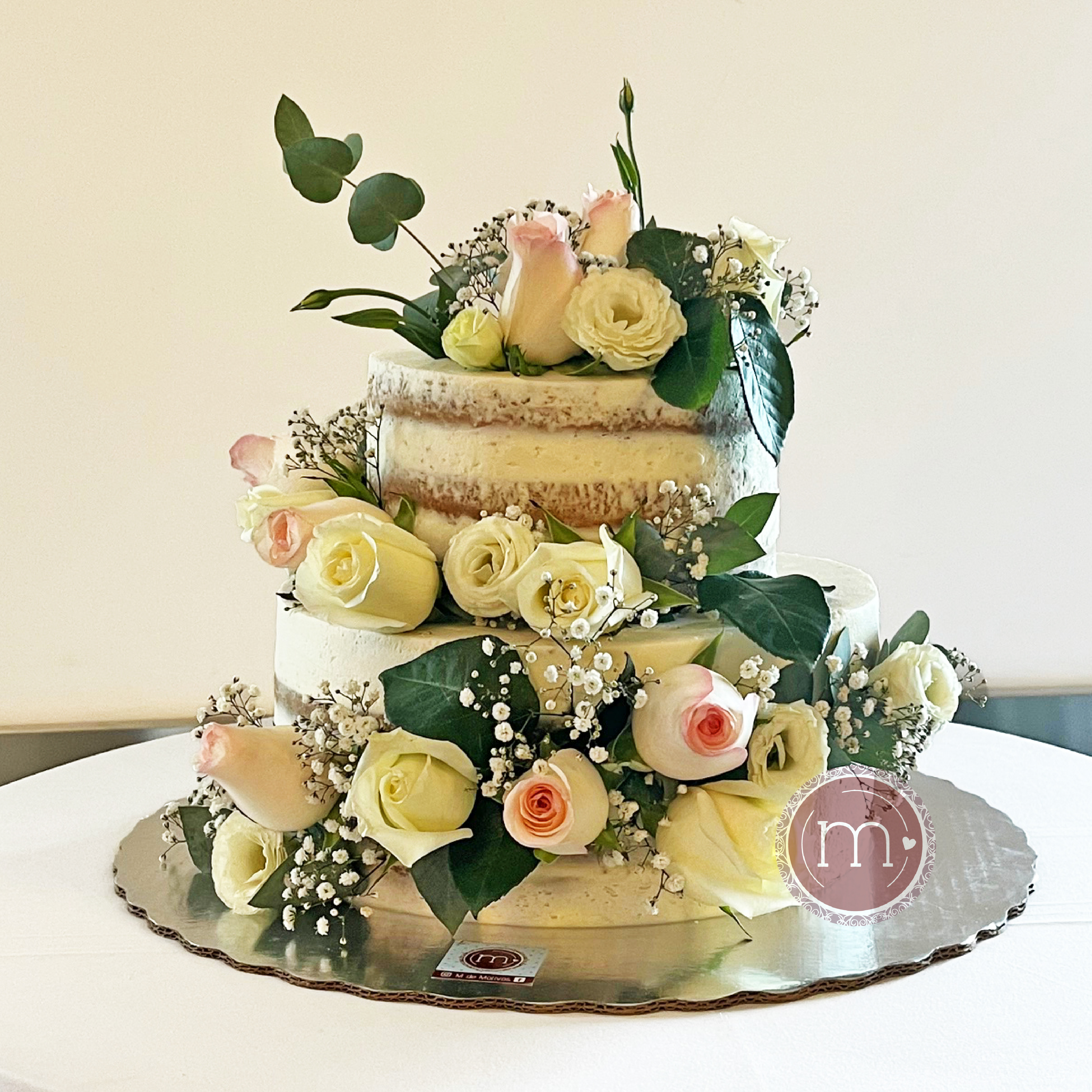 Imagen pastel con flores blancas grandes Un pastel de tres pisos con cubierta blanca, está decorado con una tira de flores, rosas blancas y hojas verdes. El pastel está sobre una base de color platado y a su vez sobre una tela blanca. Este estilo de pastel "naked cake" es un diseño ideal de pastel de bodas.