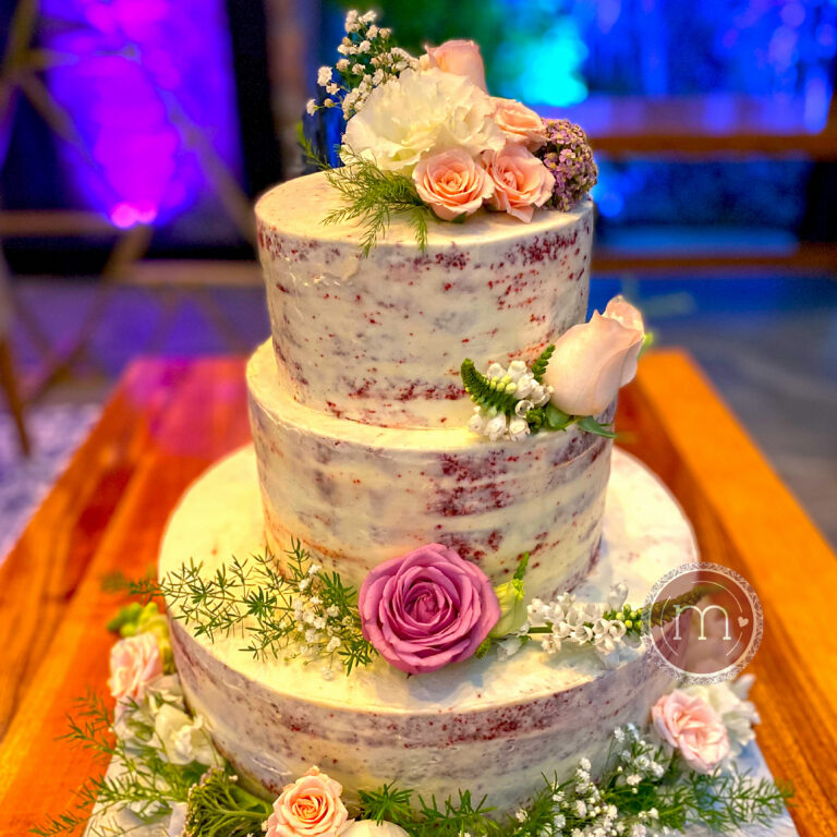 Un pastel de tres pisos red velvet estilo naked cake, decorado con rosas de color morado, rosa claro y flores baby gysophila. Este pastel se coloca sobre una mesa de madera. Un pastel de bodas de red velvet le da un toque de elegancia a una boda.