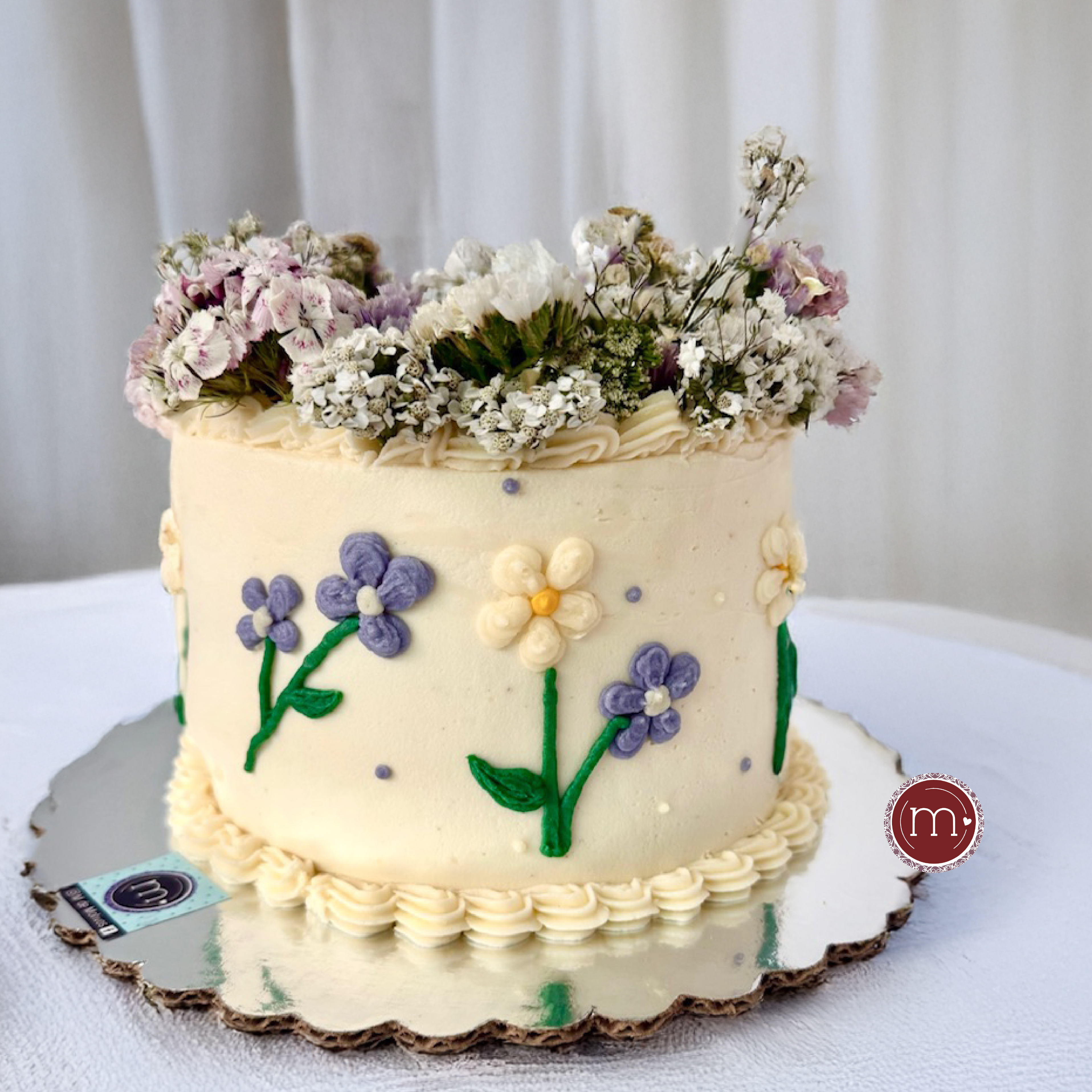 Un pastel visto desde arriba decorado con betún y blanco y flores naturales. A los lados tiene decoración de flores hechas con betún. Los pasteles personalizados y decorados con flores son los favoritos para el verano.