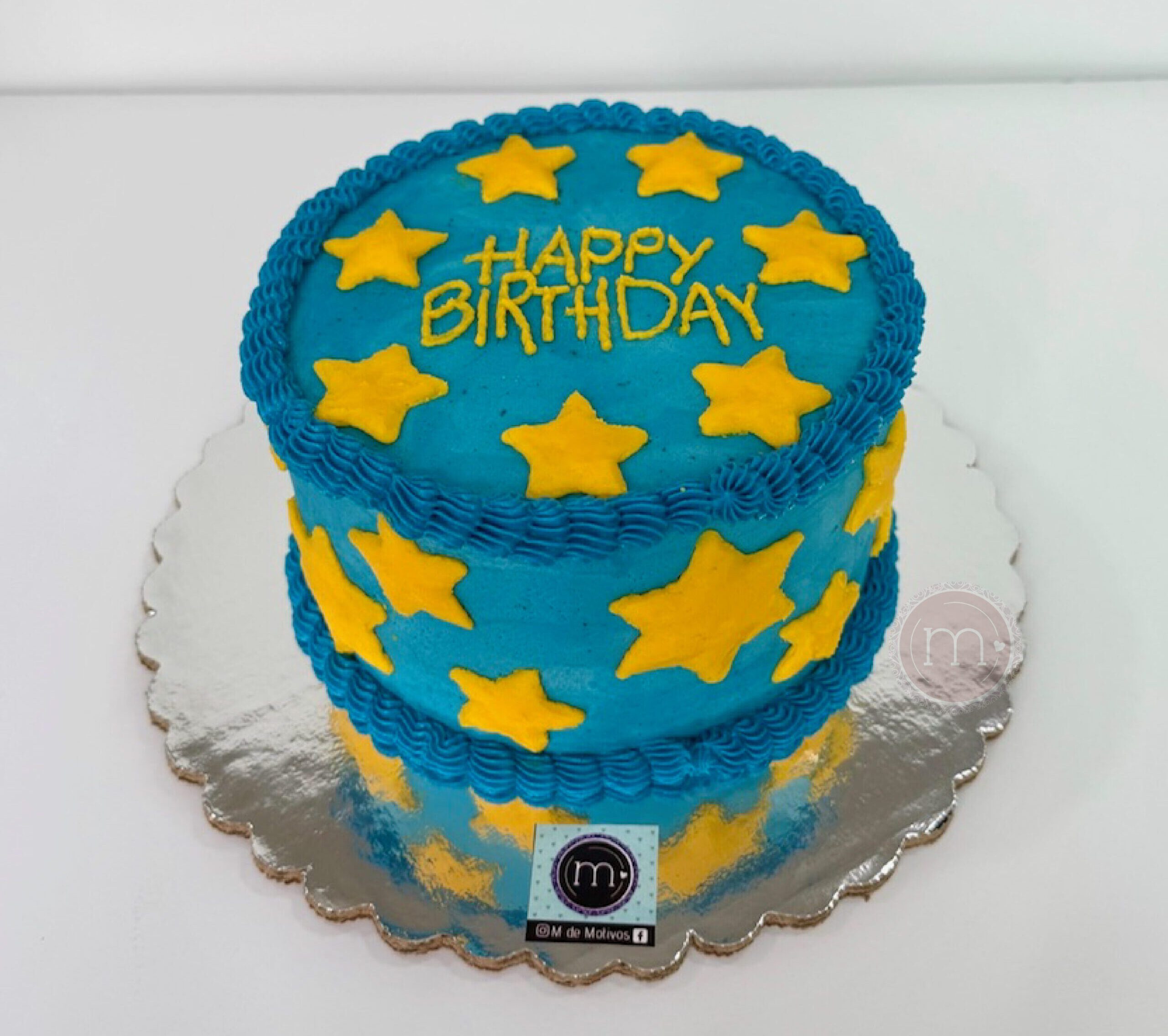 Este pastel personalizado tiene una decoración de betún color azul con estrellas amarillas. En la parte superior del pastel se escribe "Happy Birthday". El pastel se coloca sobre una base reflectante.