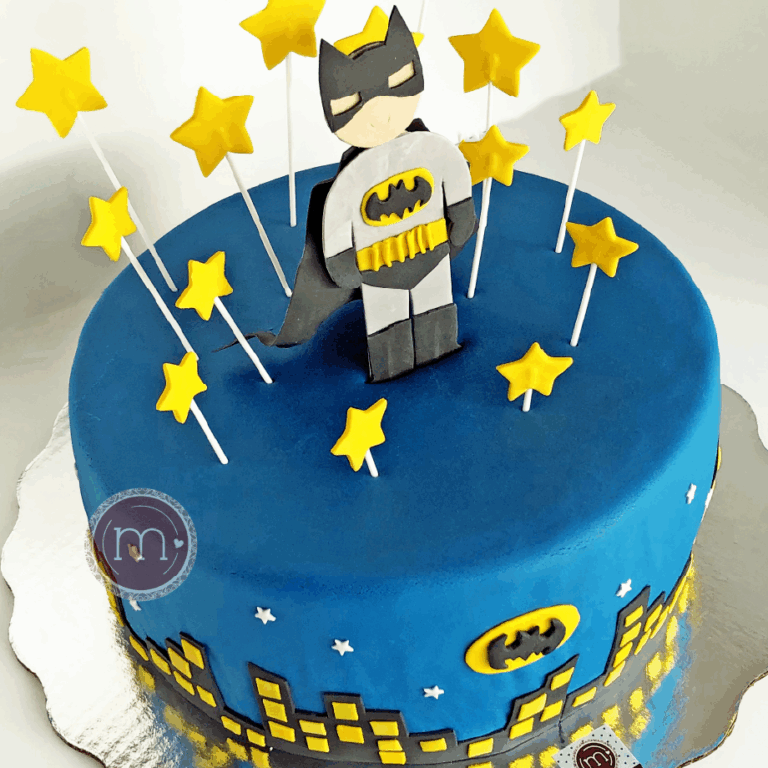 Un pastel redondo de color azul decorado con estrellas amarillas de fondant. En la parte superior se coloca una figura de fondant del personaje "Batman", Los pasteles fondant tienen colores vibrantes. El pastel se coloca sobre una base plateada reflectante.
