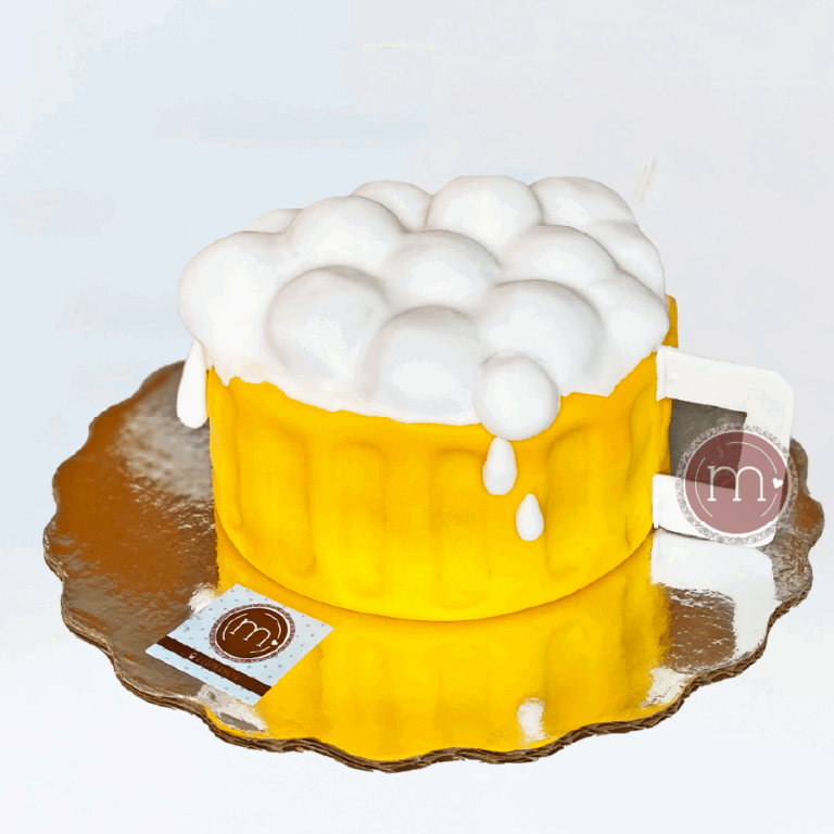 Una pastel en forma de tarro de cerveza, con una base amarilla y espuma de fondant. Los pasteles fondant son los preferidos para pasteles personalizados. El pastel se coloca sobre una base de cartón plateado reflectante. La base de cartón tiene una etiqueta con la letra M.