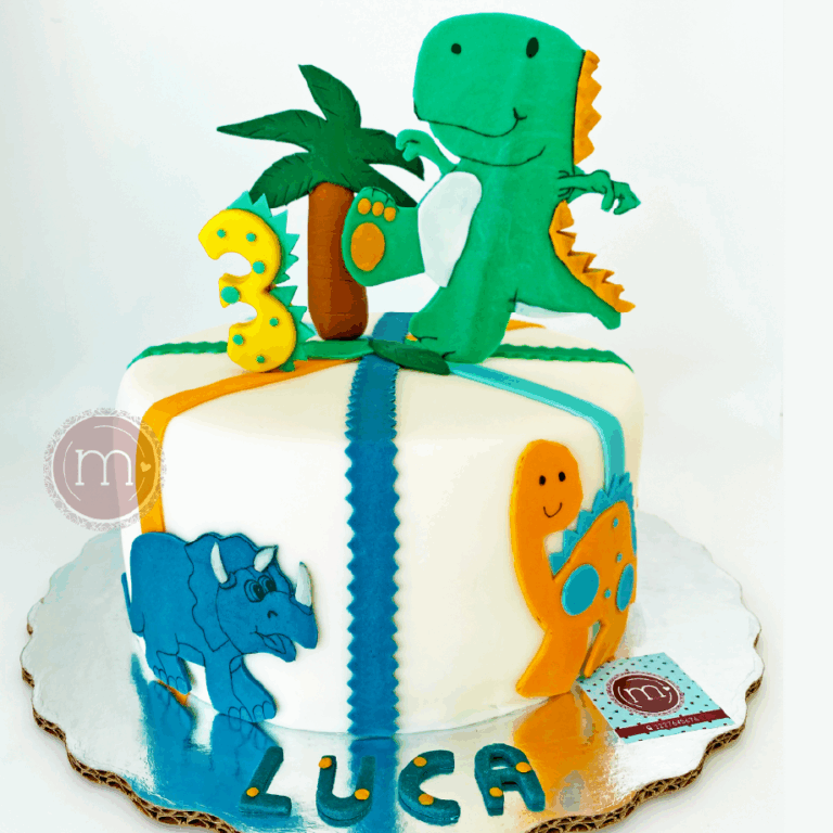 Un pastel de cumpleaños con dinosaurios decorado presenta figuras de un dinosaurio verde sonriente y una palmera en la parte superior, un número "3" amarillo y varias decoraciones de dinosaurios a los lados. En la base está escrito el nombre "LUCA" con guinda azul.