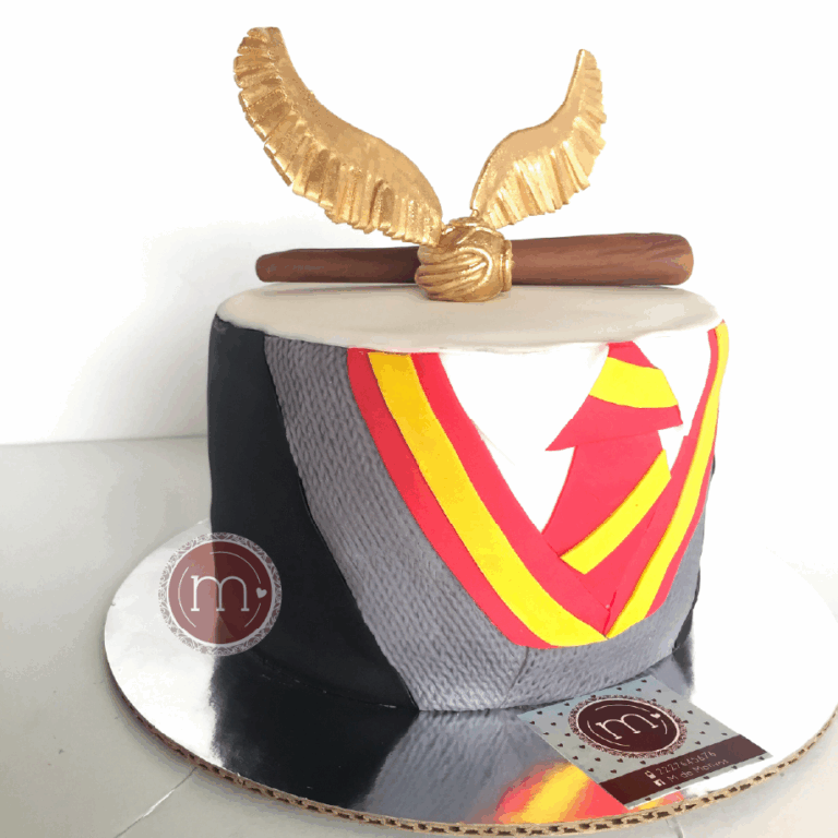 Un pastel de un piso con decoración de fondant que representa el uniforme escolar de la saga "Harry Potter". En la parte superior se coloca una "snitch dorada" de fondant y una "barita de sauco" hechas de fondant. Los pasteles fondant con temática de Harry Potter son poulares. El pastel se coloca sobre una base reflectante.