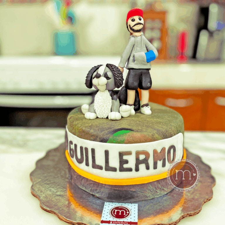Un pastel de fondant que se encuentra rodeado por una etiueta de fondan que tiene escrito en mayúsculas el nombre "GUILLERMO". En la parte superior del pastel hay un par de figuras de fondant, un perro de color gris con orejas largas y una figura de un hombre con ropa deportiva, barba y una gorra roja. El pastel se coloca sobre una base de color plateado reflejante. Los pasteles fondant personalizados son un excelente regalo de cumpleaños.