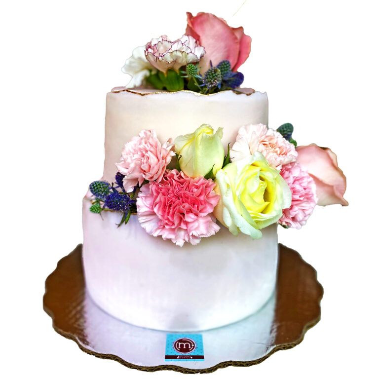 Un pastel blanco de dos pisos está decorado con flores naturales de colores rosa y morado. Los pasteles personalizados y decorados con flores son ideales para bodas o XV años. El pastel se coloca sobre una base reflectante.