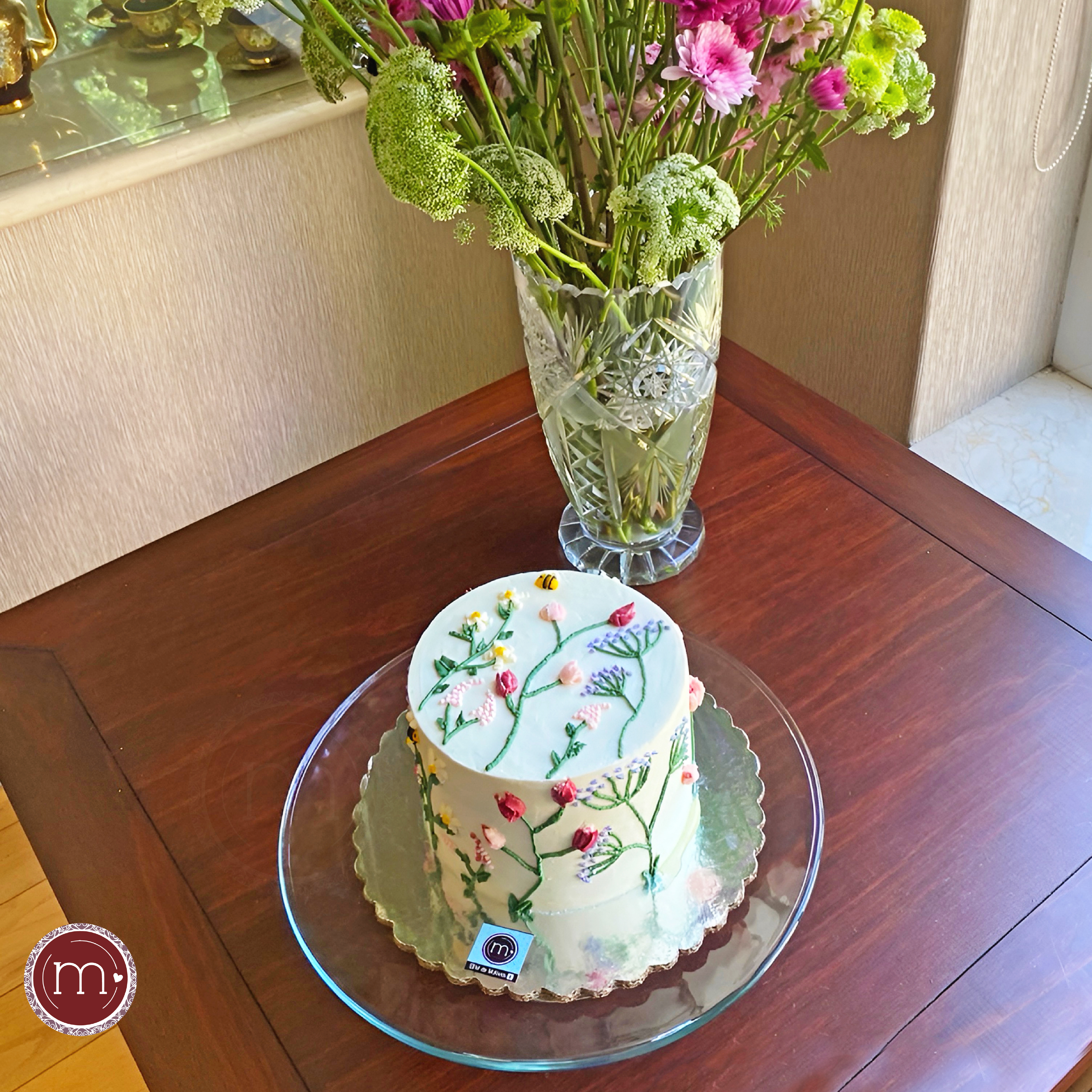 Un pastel redondo decorado con flores de betún. El pastel se coloca sobre una base de cristal y a su vez sobre una base de madera. Los pasteles personalizados se decoran de acuerdo a la temática que el cliente desee.