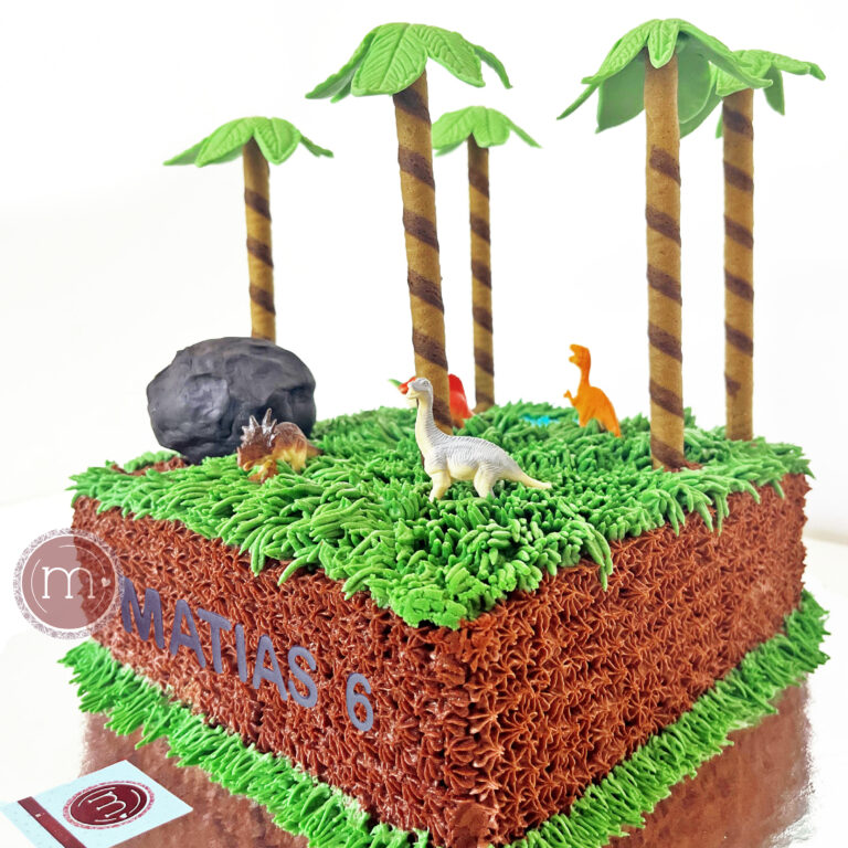 El catálogo de pasteles personalizados de repostería "m de motivos" incluye diseños como este, que es un pastel cuadrado decorado con una temática de dinosaurios, pues la superficie es un betún verde que simula el pasto. En la parte superior se colocan pequeñas figuras de dinosaurios y palmeras hechas con fondant. En la parte frontal del pastel se escribe el nombre "MATIAS 5".