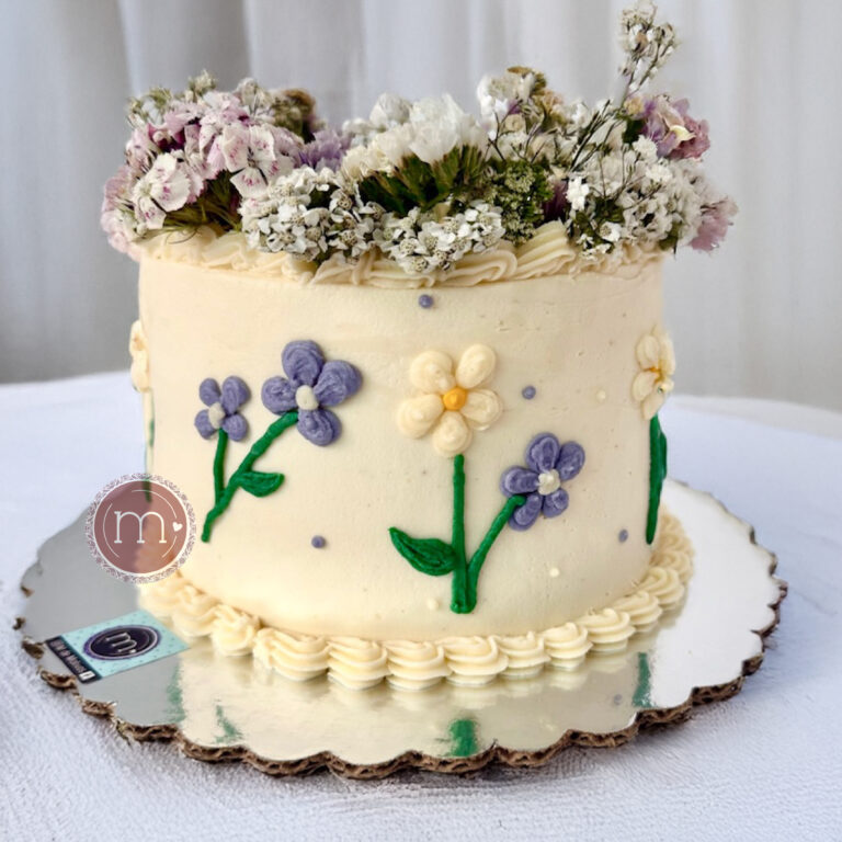 Un pastel blanco decorado con flores de betún de color morado con tallos verdes. Estos pasteles personalizados tienen detalles cuidadosamente elaborados. El pastel se coloca sobre una base reflectante.