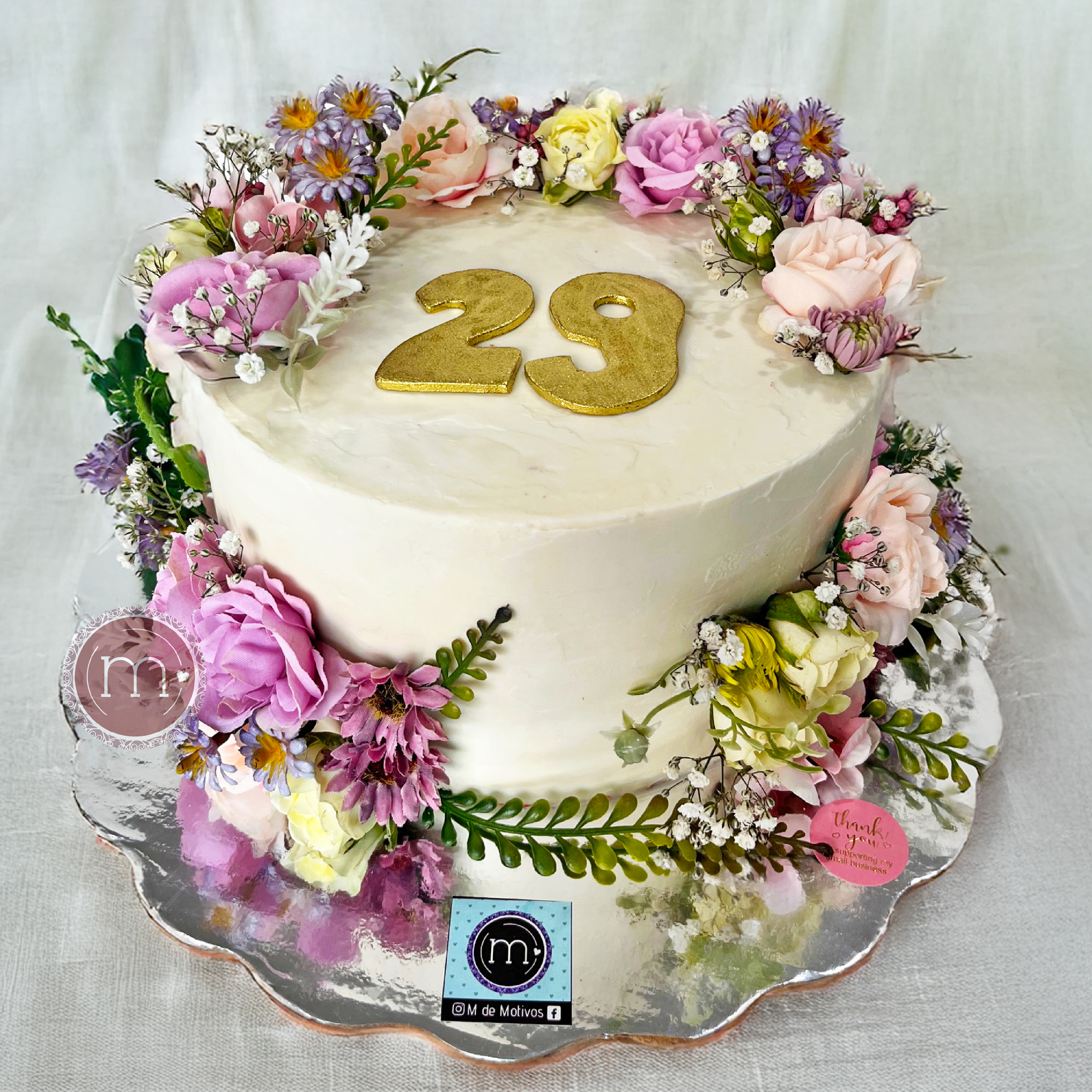 Un pastel blanco decorado con flores naturales de color morado y color rosa. Las flores van a compañadas de un follaje verde. En la parte superior se coloa un número "29" hecho de fondant. Los pasteles personalizados con flores naturales son muy populares. El pastel se coloca sobre una base reflectante.