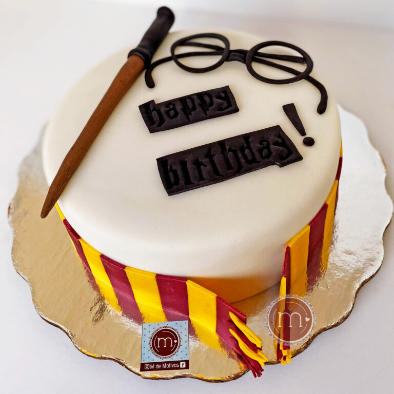 Un pastel blanco decorado con elementos e la saga de "Harry Potter", como una bufanda, unas gafas y una barita, que se colocan en la parte superior del pastel. Este tipo de decoraciones de pasteles personalizados con temáticas se suelen hacer con fondant. En la parte superior se colca un par de placas que dicen "Happy Birthday". El pastel se coloca sobre una base reflectante .