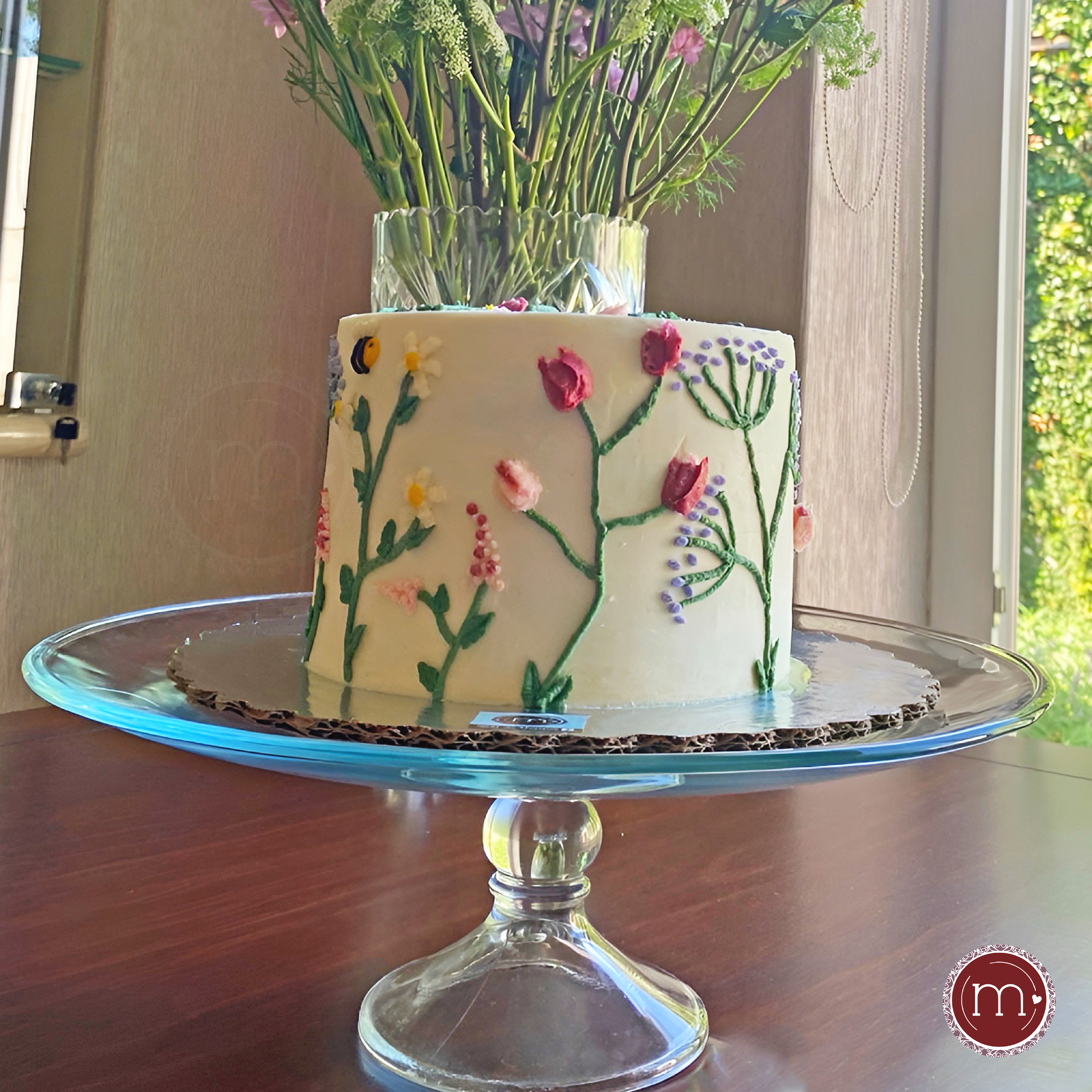 Un pastel redondo decorado con flores de betún. El pastel se coloca sobre una base de cristal y a su vez sobre una base de madera. Los pasteles personalizados se decoran de acuerdo a la temática que el cliente desee.