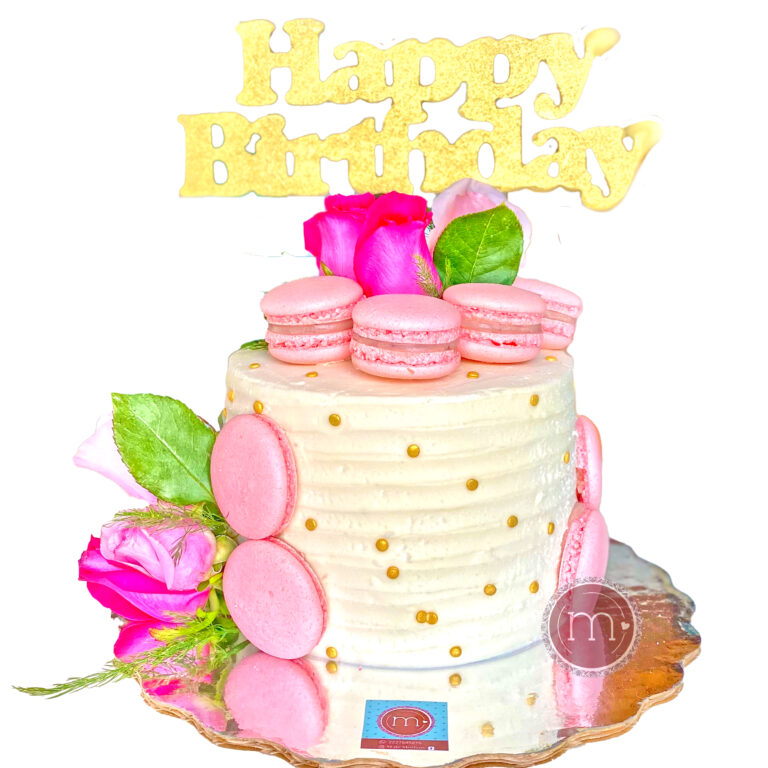 Un pastel blanco decorado con perlas doradas y macarons rosados. En la parte superior se coloca una rosa y un letrero que dice "Happy Birthday". En repostería m de motivos hacemos pasteles personalizados para cumpleaños. El pastel se coloca sobre una base reflectante.
