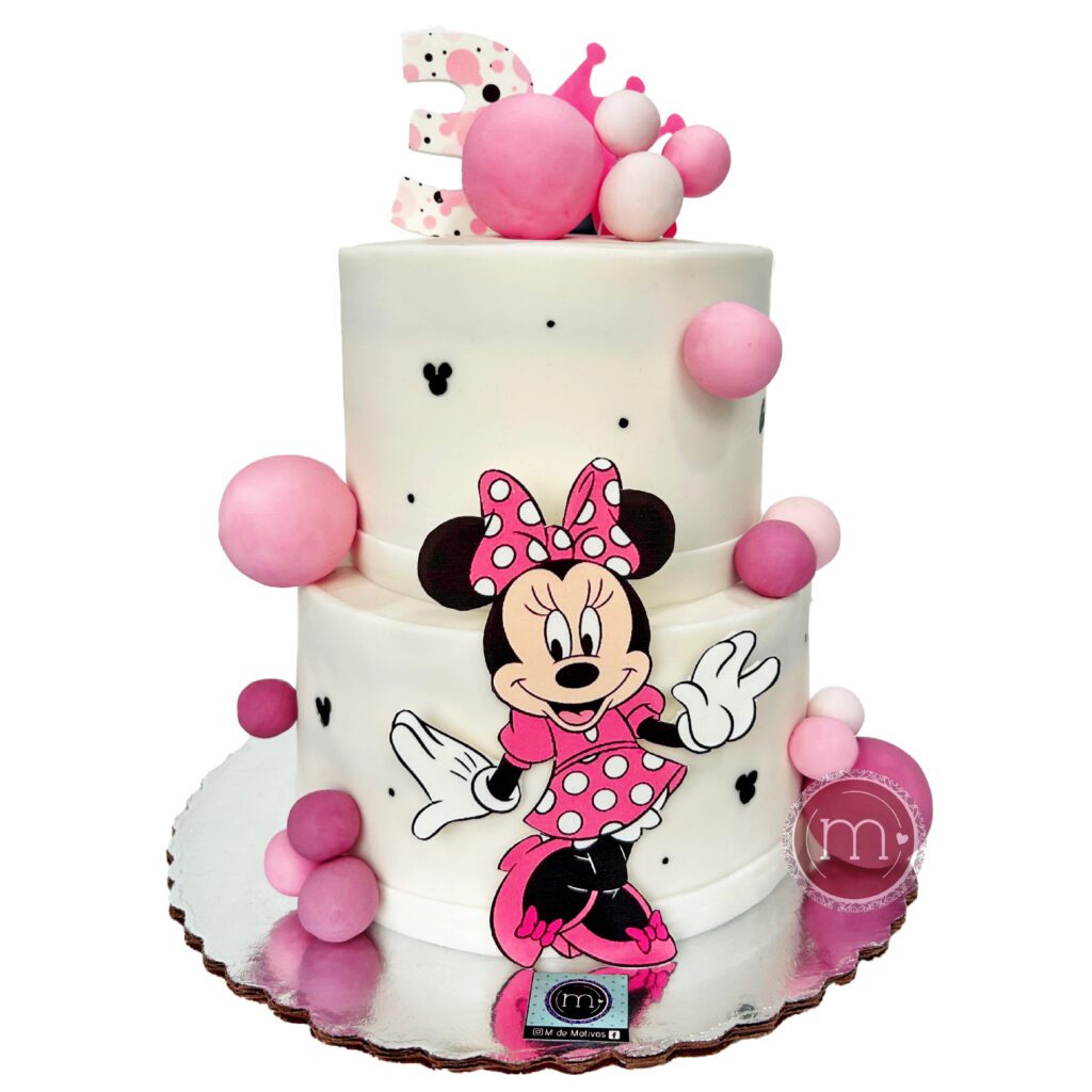 Un pastel de dos pisos de color blanco tiene decoraciones con temática del personaje "Minnie" En la repostería "M de motivos" se hacen pasteles personalizados con temáticas. El pastel se coloca sobre una base reflectante.