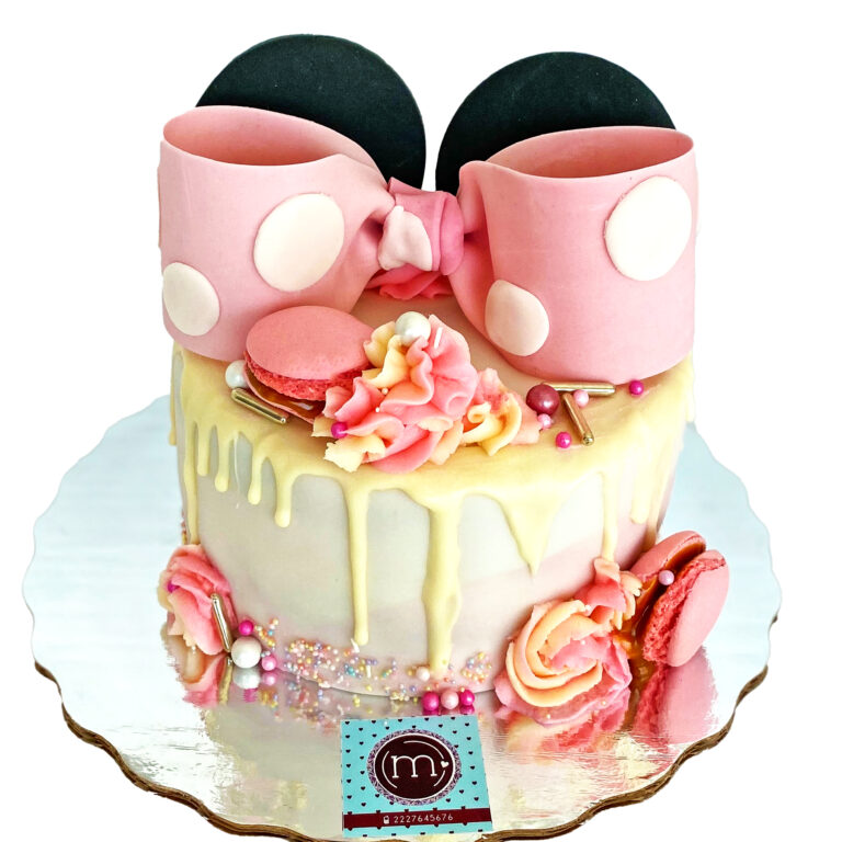 Un pastel decorado con macarrones de color rosa, sprinkles en tonos rosados y una cubierta de chocolate blanco que escucrre desde la parte superior. Encima se coloca un moño rosado de fondant con unas orejas de fondant similares a las del personaje de "Minnie". Estos pasteles personalizados son ideales para fiestas temáticas. El pastel se coloca sobre una base reflectante.