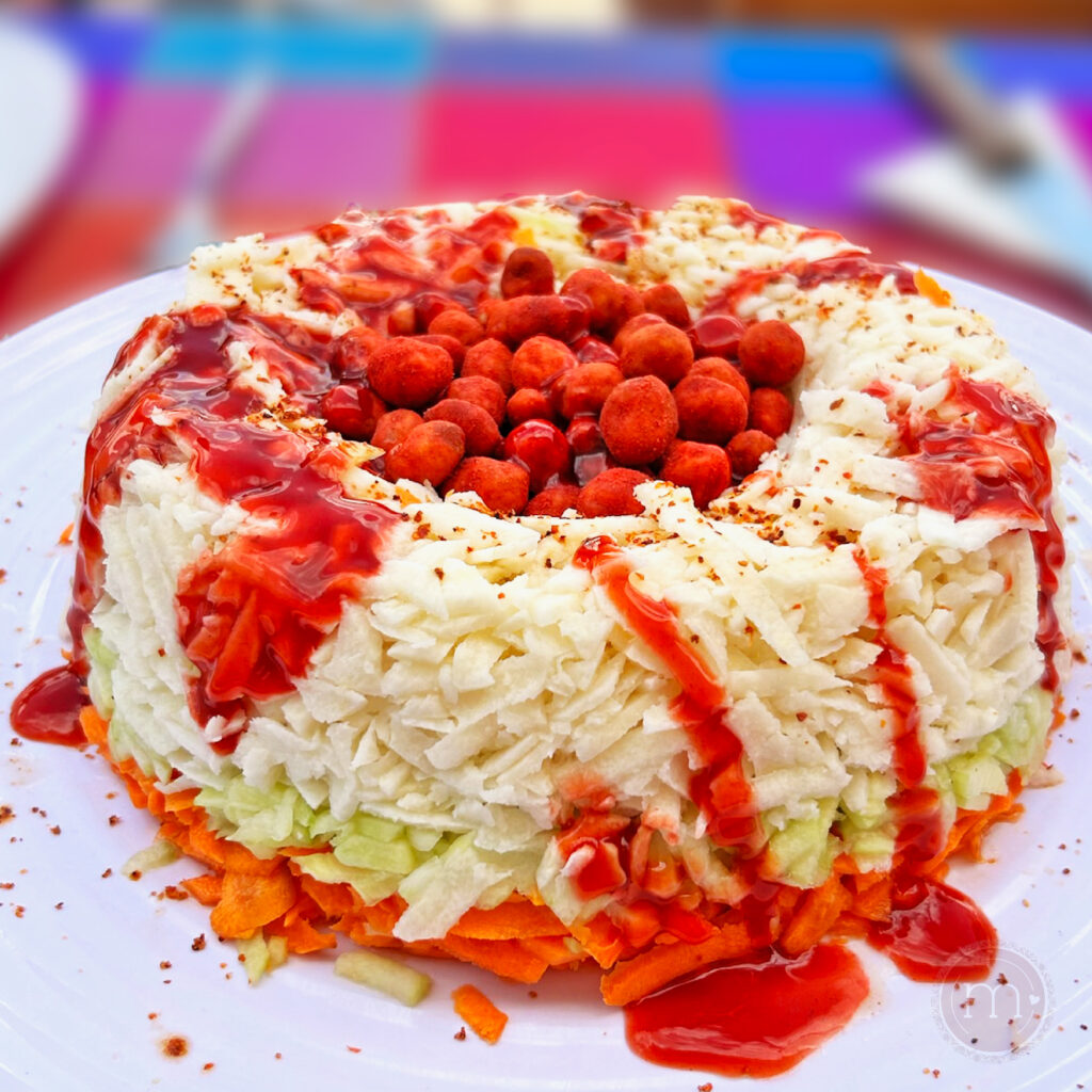 Rosca de botana para la noche mexicana. L rosca es de zanahoria con pepino y jícama. En el centro tiene cacahuates picosos y está bañada con salsa de chamoy. La rosca se coloca sobre un plato blanco.