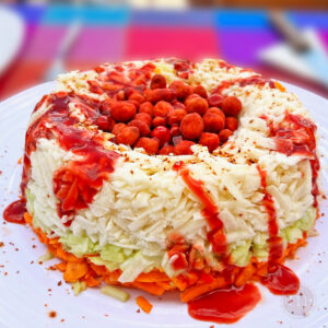 Rosca de botana para la noche mexicana. L rosca es de zanahoria con pepino y jícama. En el centro tiene cacahuates picosos y está bañada con salsa de chamoy. La rosca se coloca sobre un plato blanco.