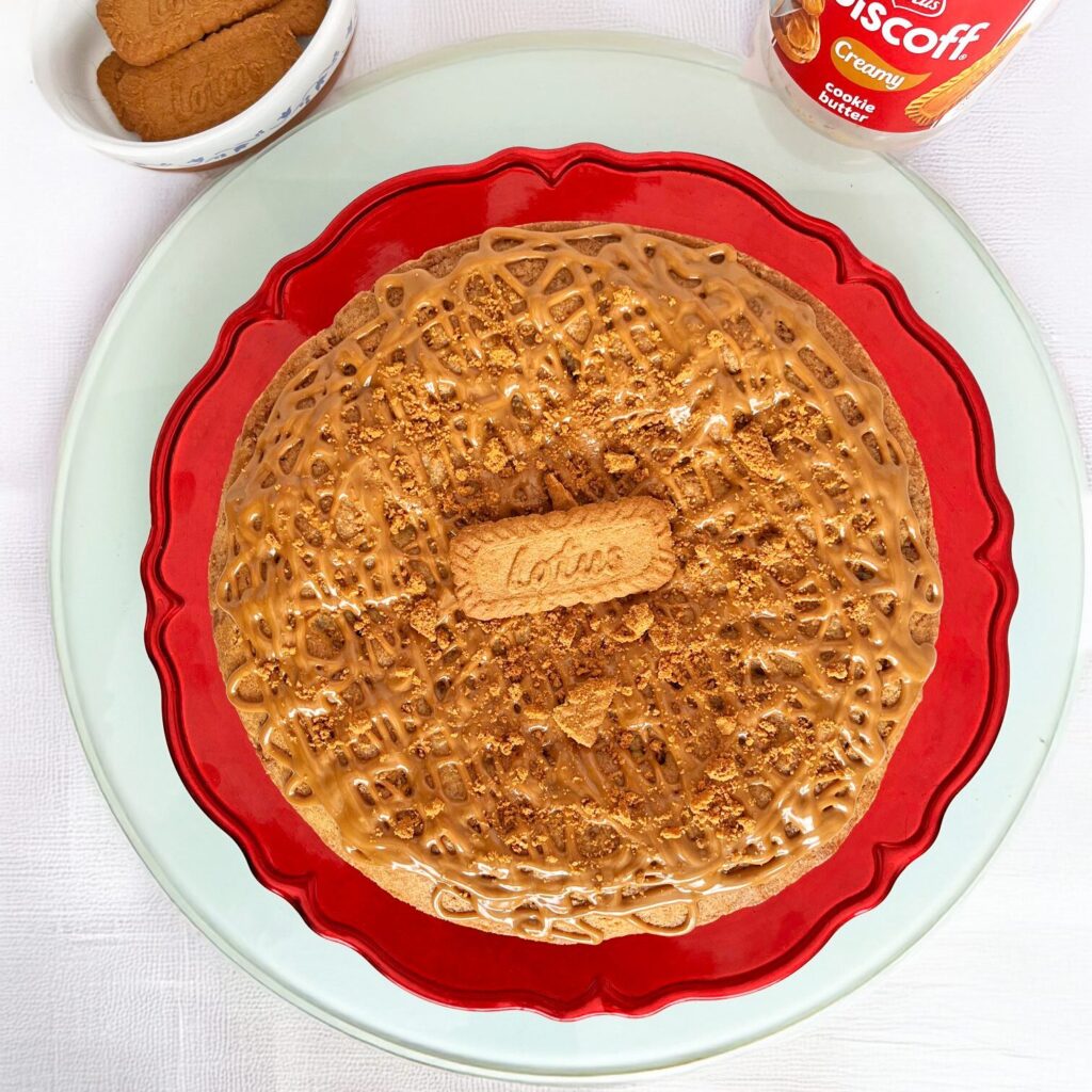 Un pastel de galleta Lotus decorado con crema Biscoff en la parte superior y con un galleta Lotus en el centro. Los postres de otoño de m de motivos son la opción ideal para las reuniones de Halloween. El pastel se coloca sobre una base de color rojo y sobre una base blanca. Lo acompañan un envase de crem Biscoff y un recipiente con galletas.