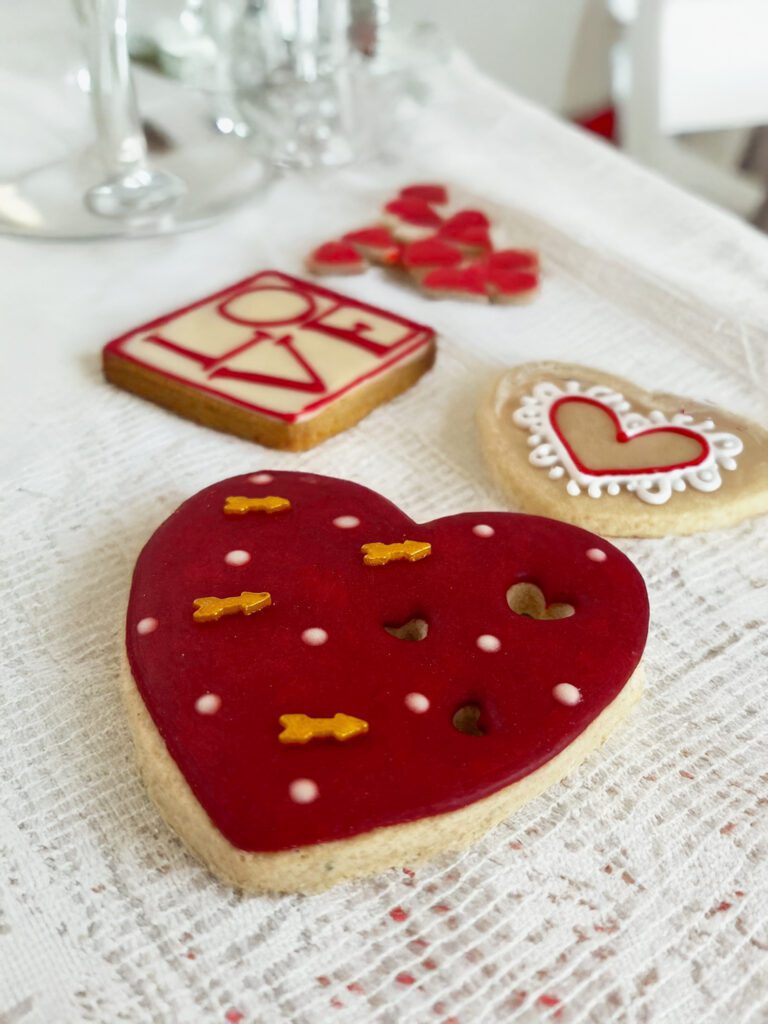 Una galleta en forma de corazón decorada con glaseado rojo. Al fondo se ve una galleta cuadrada que con glaseado tiene escrito "LOVE" y galletas mini en forma de corazón. Si buscas ideas para regalar en San Valentin, en m de motivos repostería poblana aceptan galletas personalizadas. Las galletas se colocan sobre un mantel de tela blanco.
