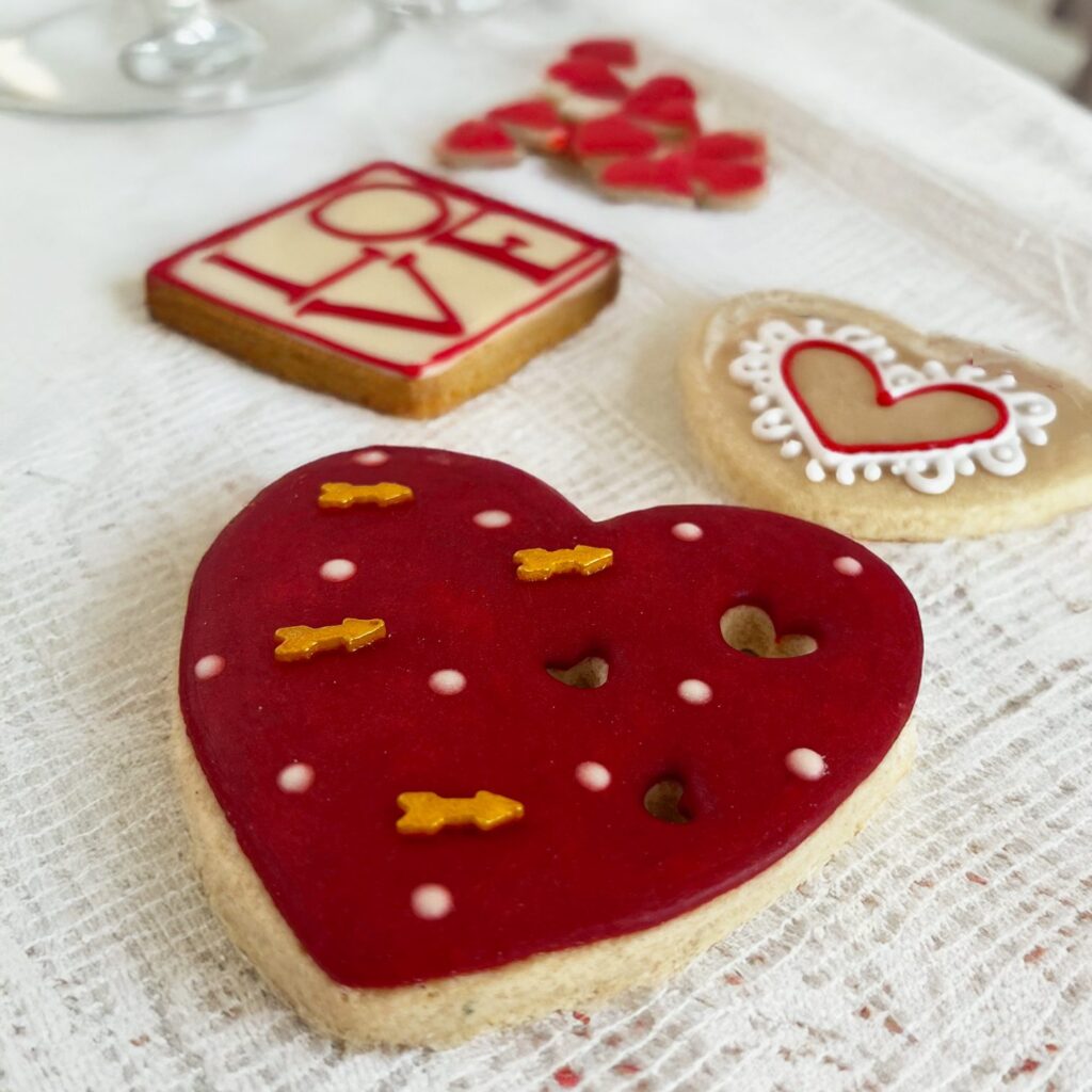 Una galleta en forma de corazón decorada con glaseado rojo. Al fondo se ve una galleta cuadrada que con glaseado tiene escrito "LOVE" y galletas mini en forma de corazón. Si buscas ideas para regalar en San Valentin, en m de motivos repostería poblana aceptan galletas personalizadas. Las galletas se colocan sobre un mantel de tela blanco.