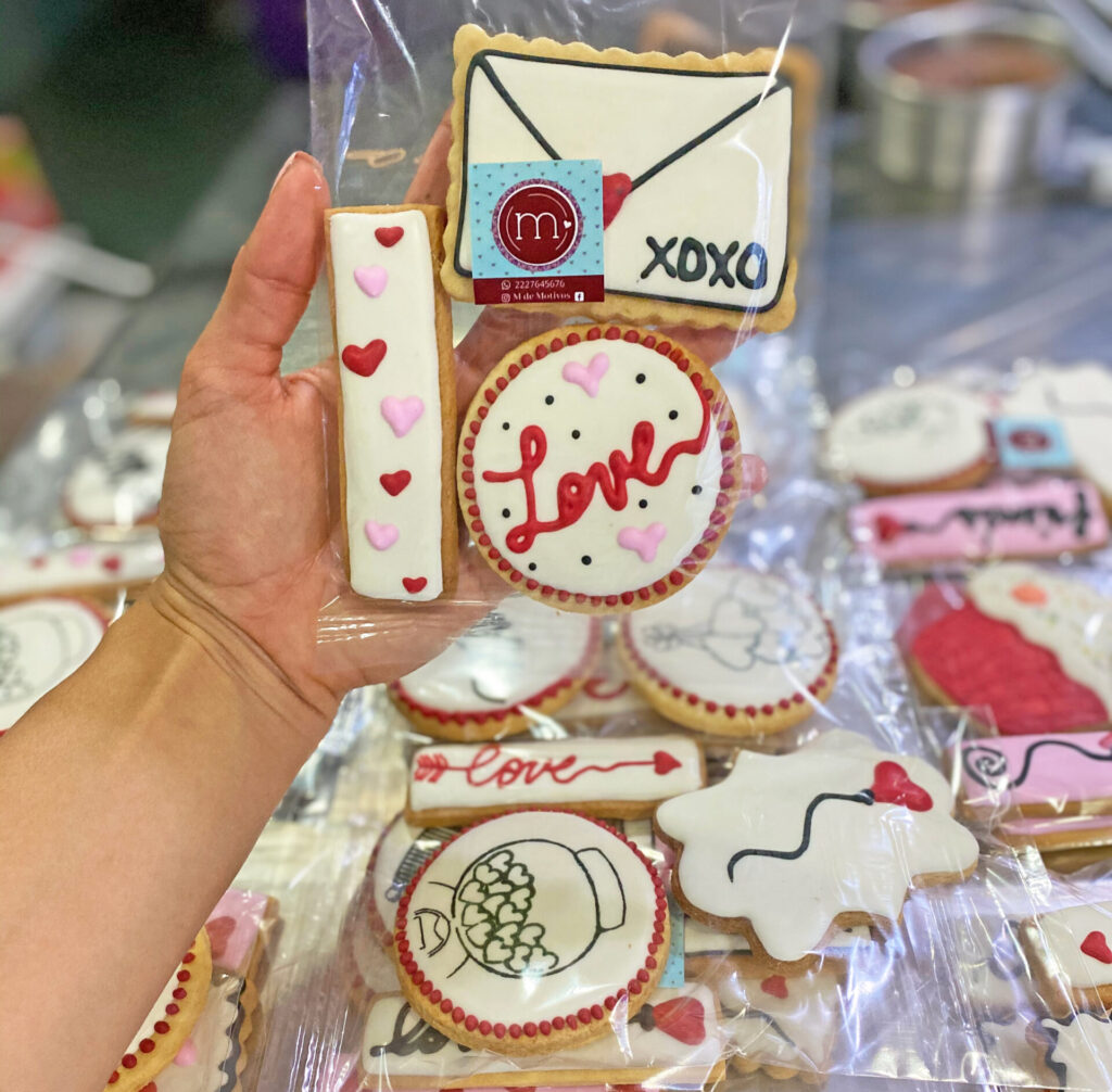 Una mano sostiene una bolsa transparente con 3 galletas decoradas con glaseado blanco. Estas ideas para san valentin tienen mensajes como la palabra "Love" o "XOXO". Al fondo se observan más galletas decoradas.