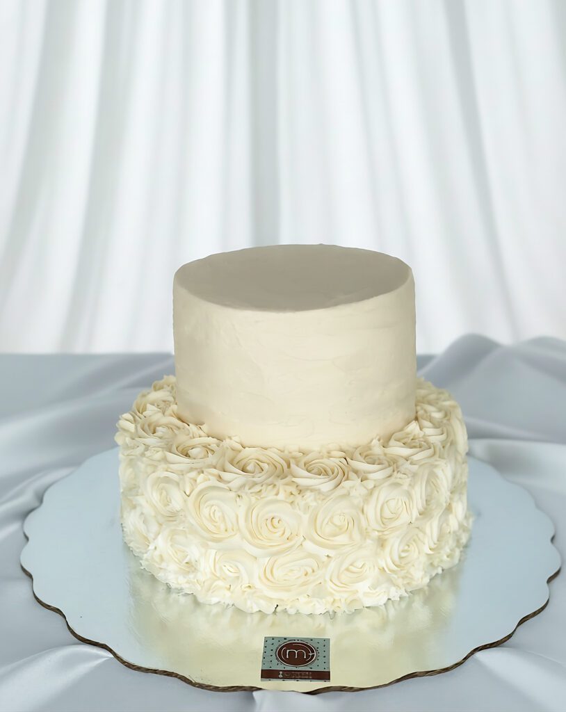 Un pastel de dos pisos color blanco decorado con betún y rosetones de betún blanco en el piso inferior. Los pasteles de boda sencillos tinenen decoraciones pequeñas y muy poco color. Este pastel se coloca sobre una base de un mantel de tela color blanco.