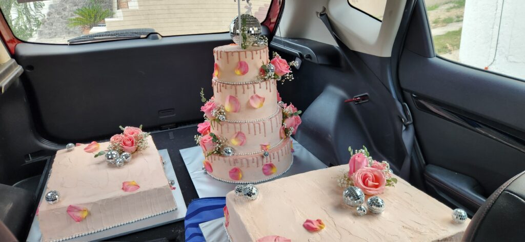 M de motivos, pasteles personalizados Puebla transporta 3 pasteles en la cajuela de un auto. Dos pasteles son planchas de color rosa decoradas con flores naturales. En medio se coloca un pastel de 4 pisos de color rosa con flores naturales y bolas tipo disco.