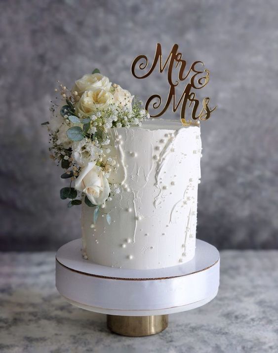 Un pastel de boda color blanco, decorado con perlas, flores blancas y un topper. Ideas de pasteles decorados con flores para bodas peuqueñas en Puebla. El pastel se coloca sobre una base blanca.
